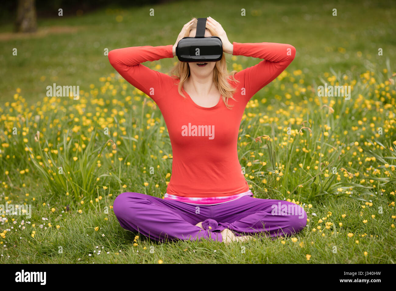 Una donna a praticare yoga nel parco mentre wering una realtà virtuale auricolare. Foto Stock