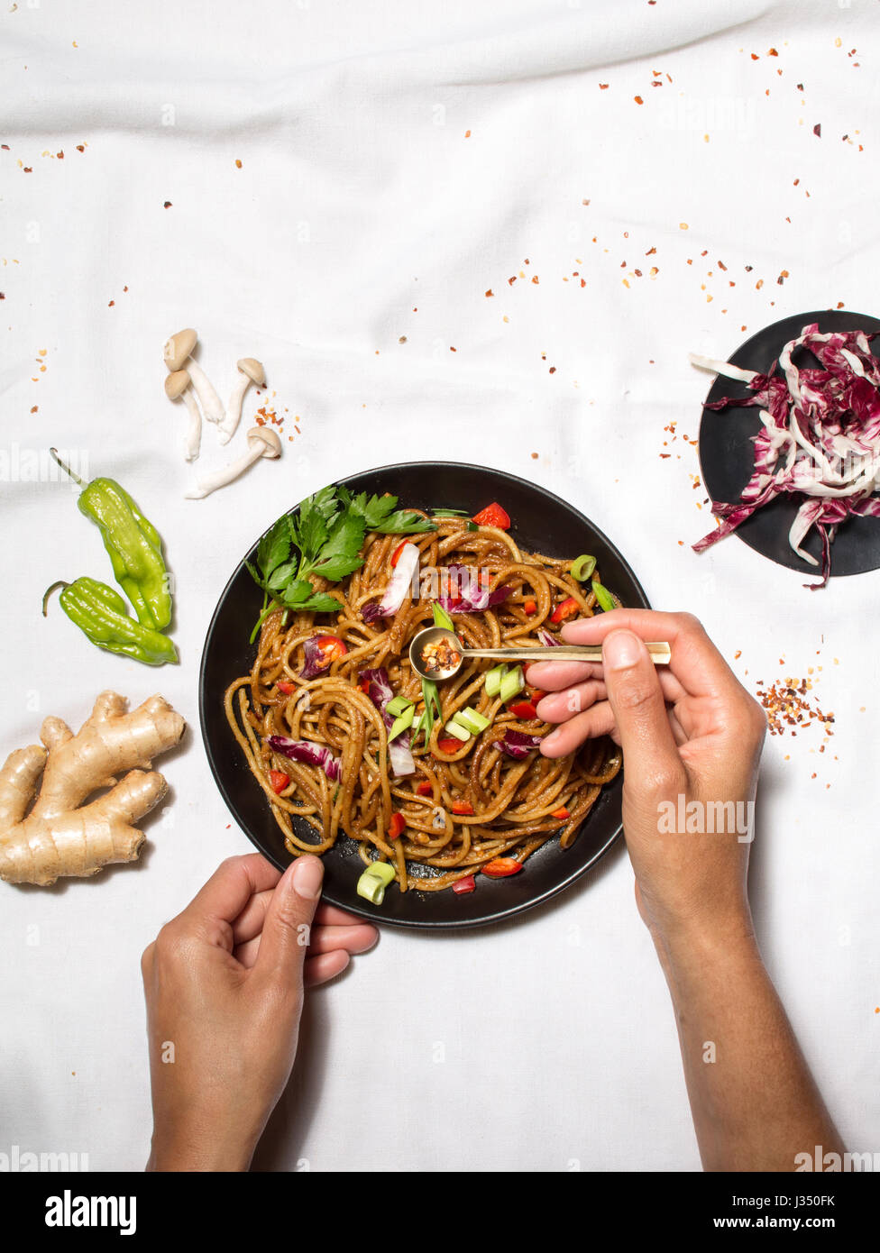 Scatto dall'alto di una donna con le mani in mano dando il tocco finale a un noodle STIR FRY, con ingredienti aggiuntivi su un tavolo bianco panno Foto Stock