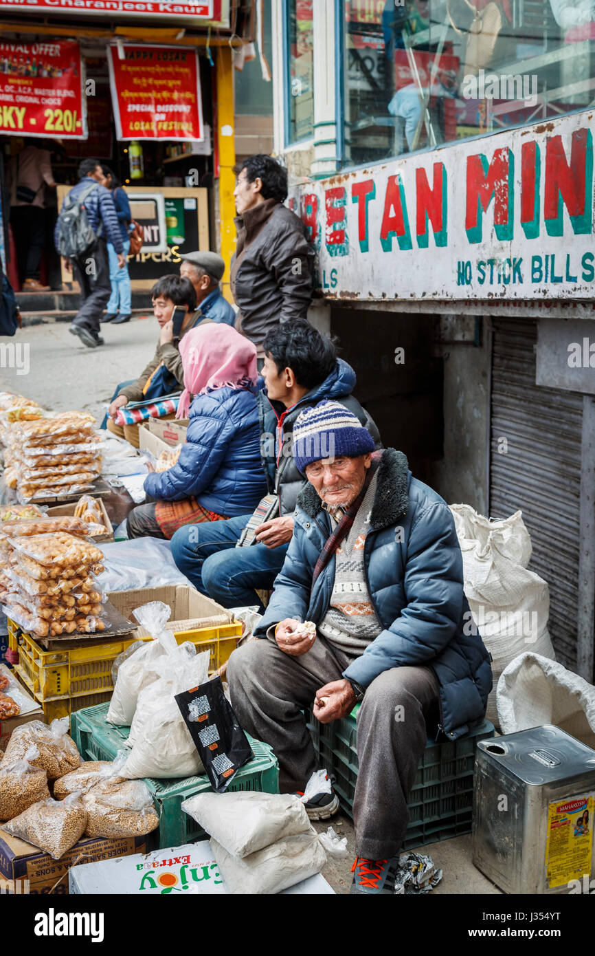 Strada locale venditore, un vecchio uomo di etnia tibetana, seduti a mangiare una banchina in stallo McLeodGanj, Dharamshala, Himachal Pradesh, India del nord Foto Stock