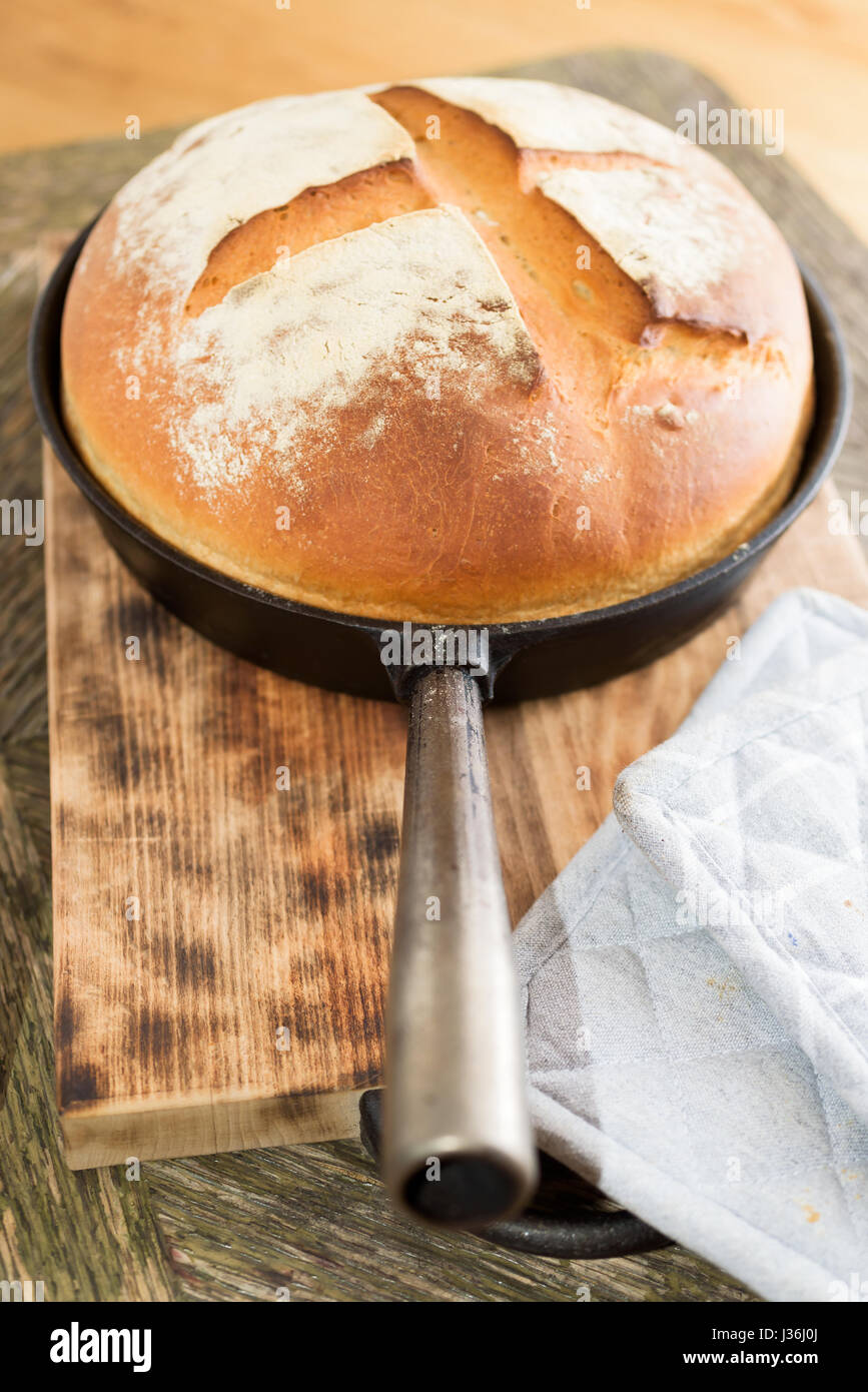 Appena cotto il pane artigianale in padella in ghisa sul tagliere di legno. Potholder grigio a lato. Foto Stock