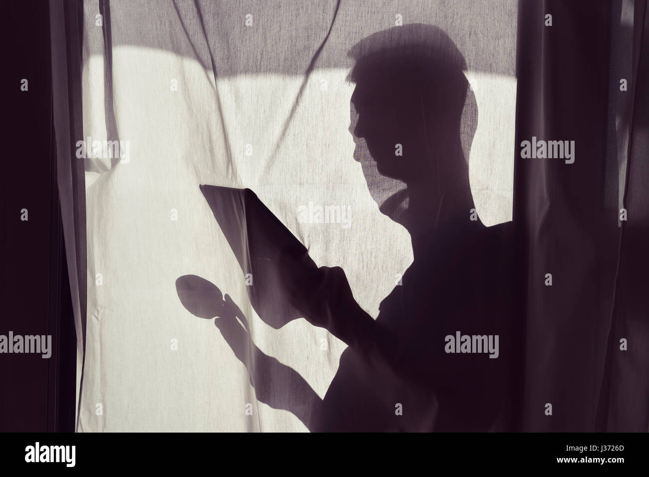 La silhouette di un giovane uomo dietro una tenda bianca con una tazza di caffè o di tè in una mano e una compressa nell'altra mano Foto Stock