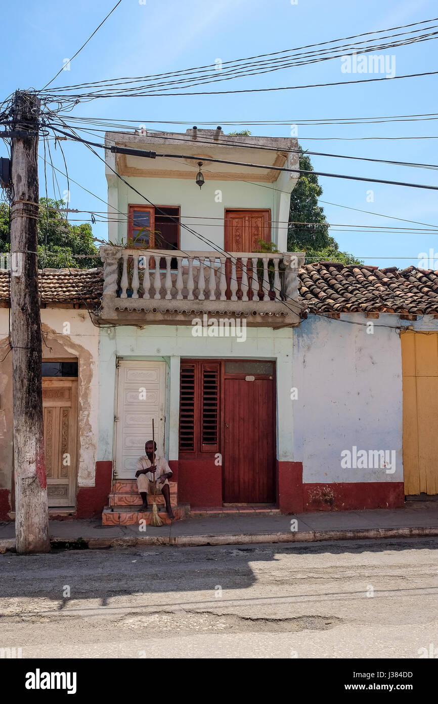 Scena di strada in Trinidad, Sancti Spiritus, Cuba. Cubano locale uomo seduto di fronte al suo sbriciolamento home azienda una scaletta di legno. Foto Stock