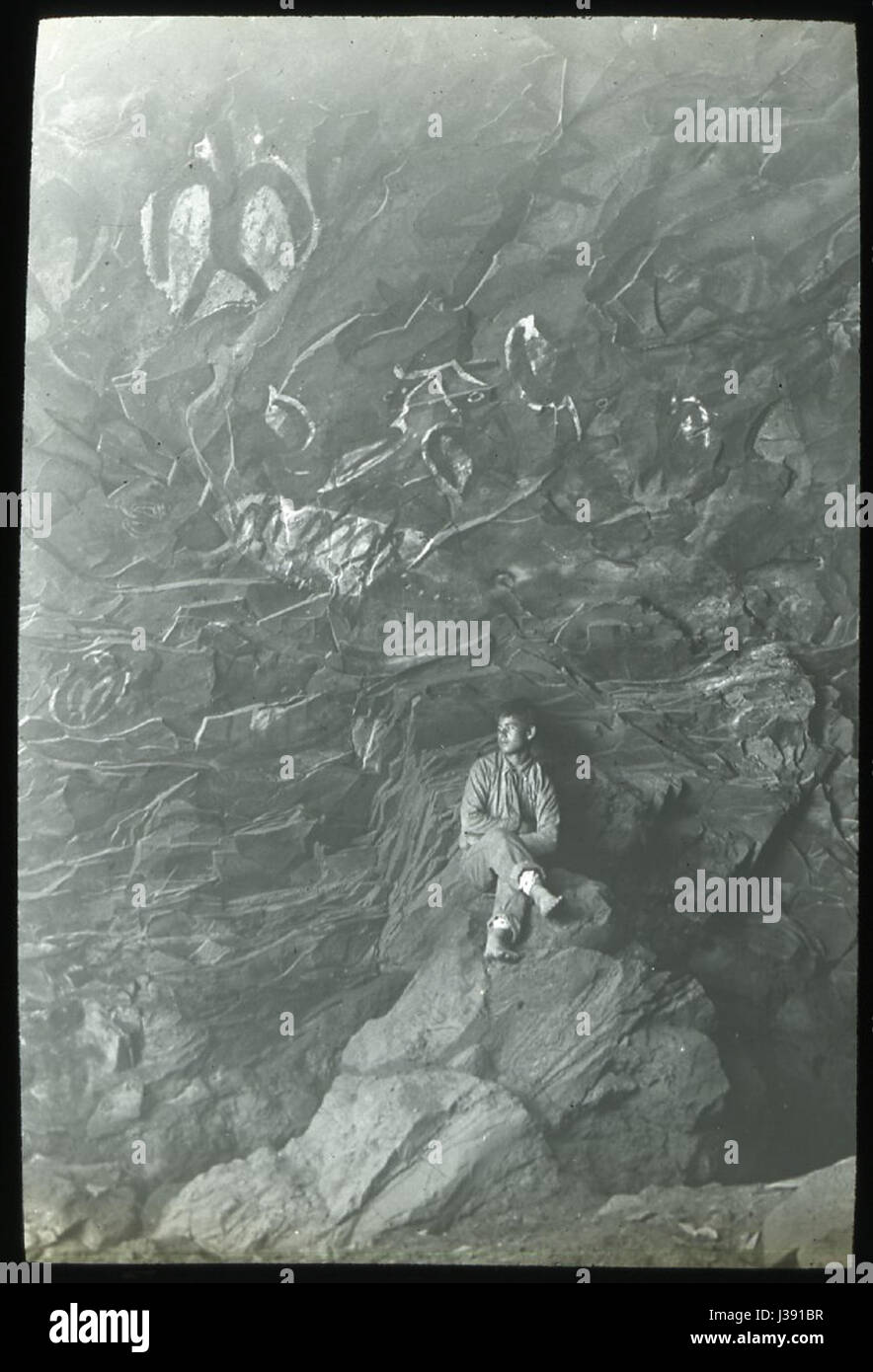 Questa immagine raffigura un ragazzo cileno seduto su una roccia all'interno di una grotta con arte rupestre dipinta in colori bianchi e scuri. L'opera fa parte della spedizione Mana sull'Isola di Pasqua, raccolta dal British Museum. Le pitture rupestri riflettono le tradizioni artistiche delle culture indigene della regione. Foto Stock