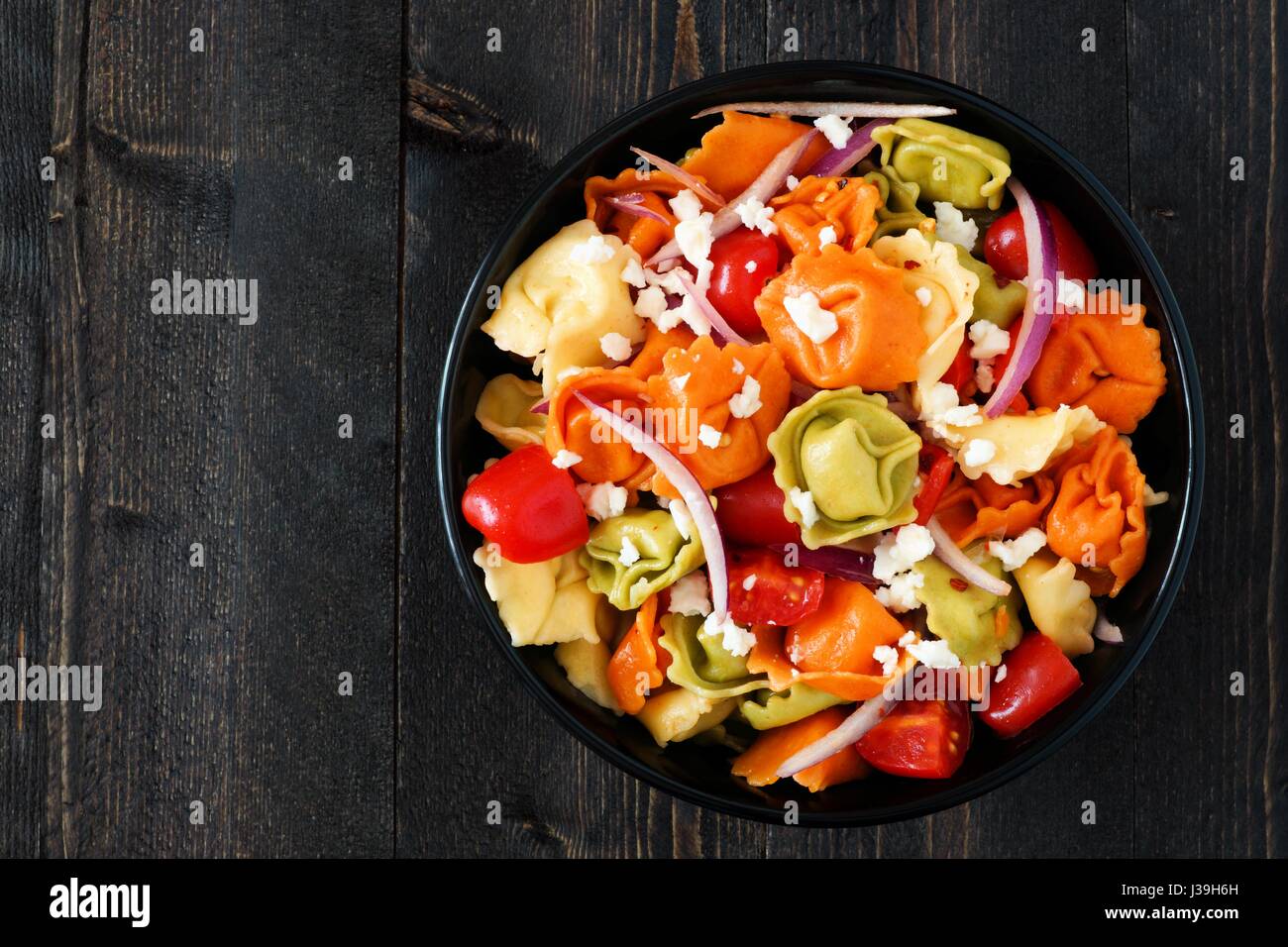 Ciotola multicolore di tortellini con insalata di pomodoro e cipolle, vista aerea su un legno scuro dello sfondo Foto Stock