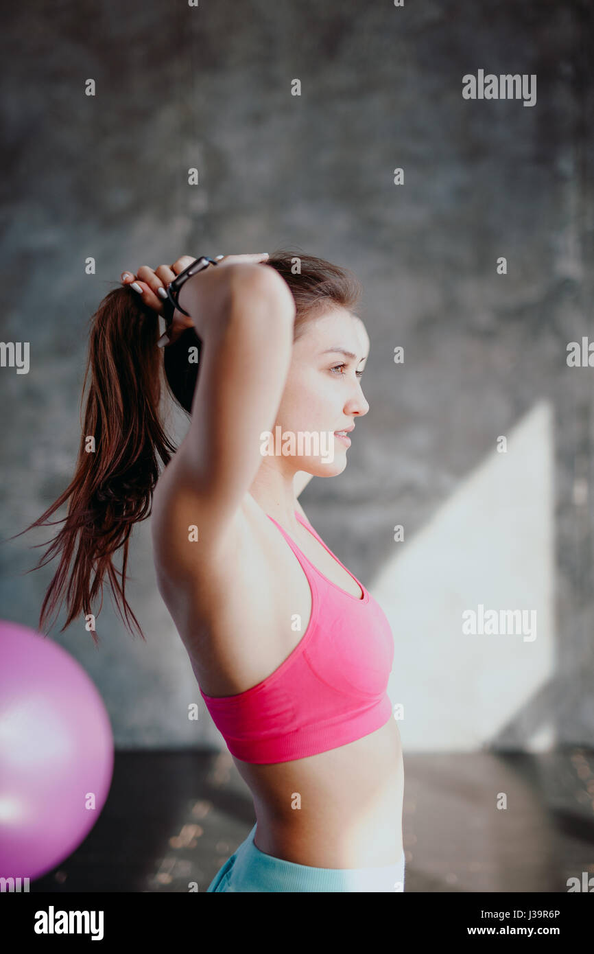 Womans break in allenamento per legare i capelli. Atleta femminile capelli di legatura prima del suo allenamento con i pesi. Bella bruna seduto alla palestra con asciugamano rosso Foto Stock