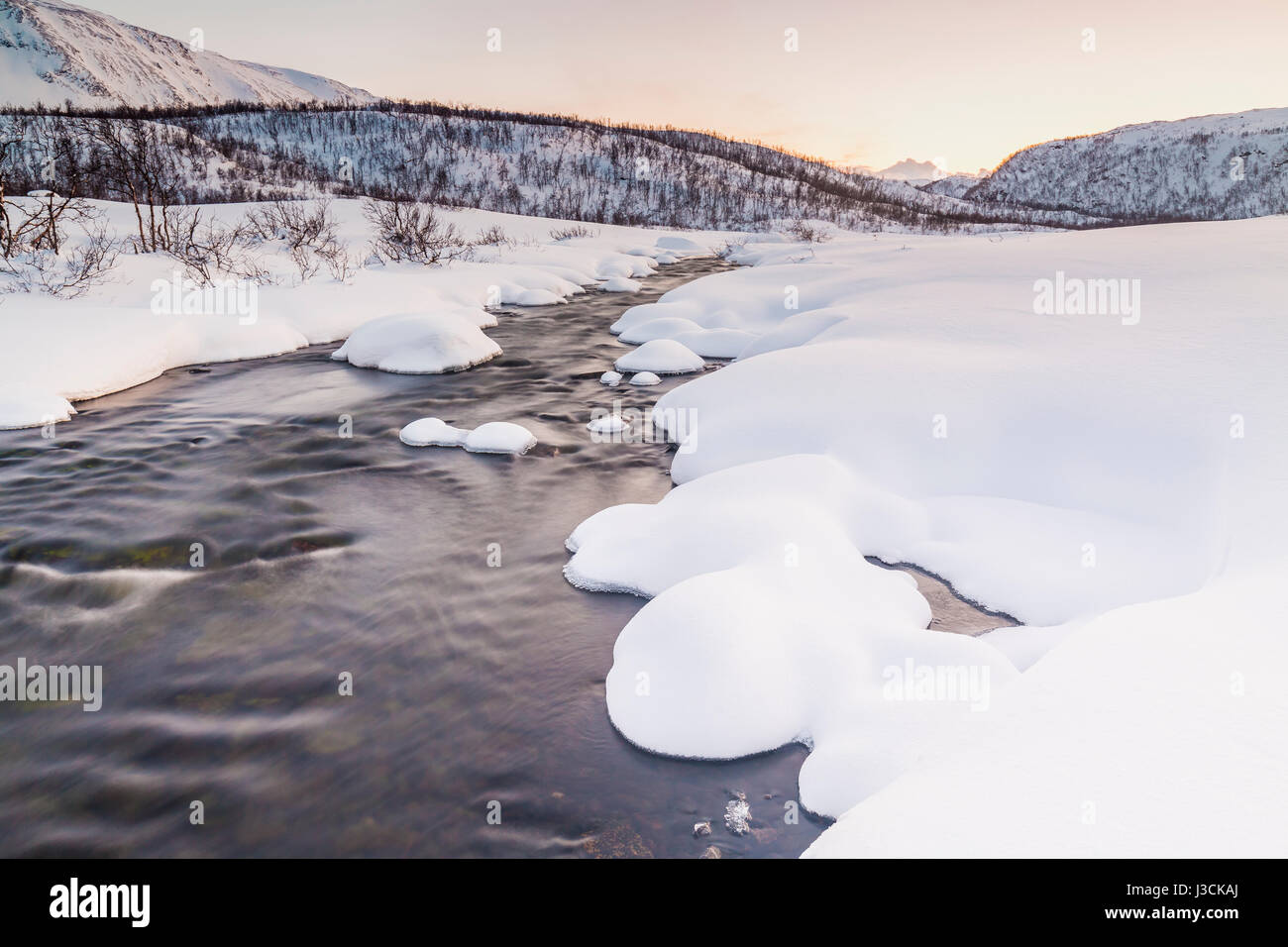 Un piccolo flusso di acqua con rocce in primo piano con un artico, paesaggio innevato in background. Il cielo è chiaro, multiple e vibrante col Foto Stock