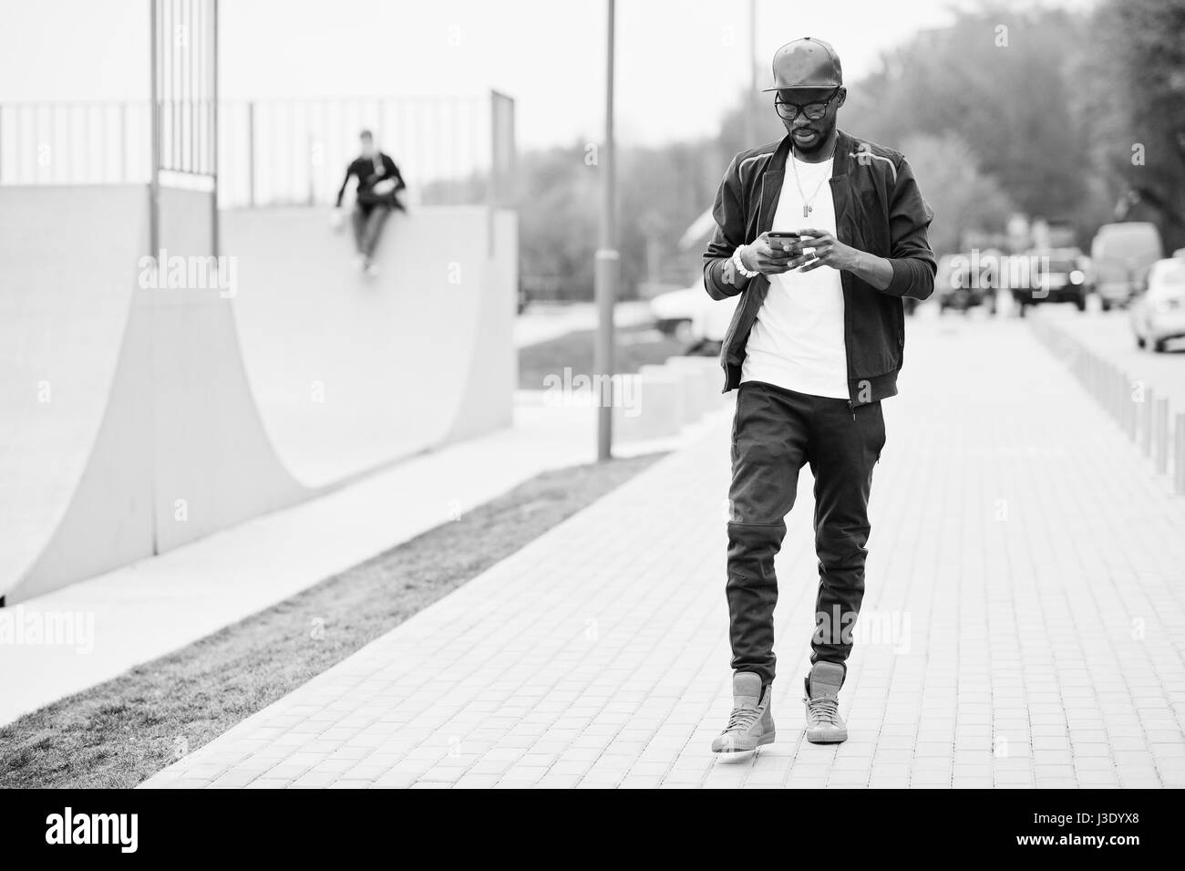 Ritratto di stile americano africano uomo su sportswear, cappello e occhiali cercando sul suo telefono cellulare. Gli uomini neri modello a piedi a skate park. Foto Stock