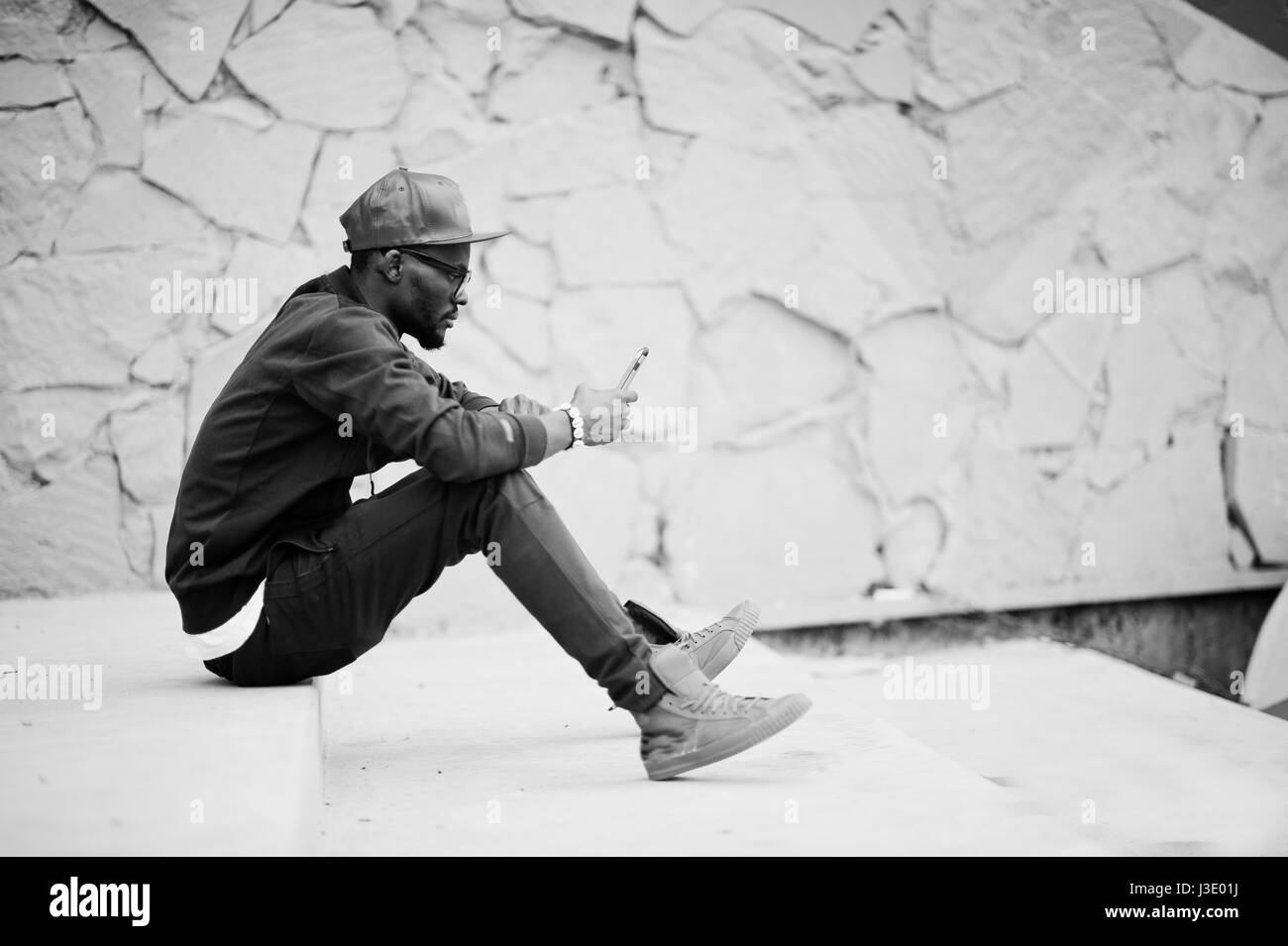 Ritratto di stile americano africano uomo su sportswear, cappello e occhiali seduti sulle scale con il telefono a portata di mano. Gli uomini neri modello street fashion. Foto Stock