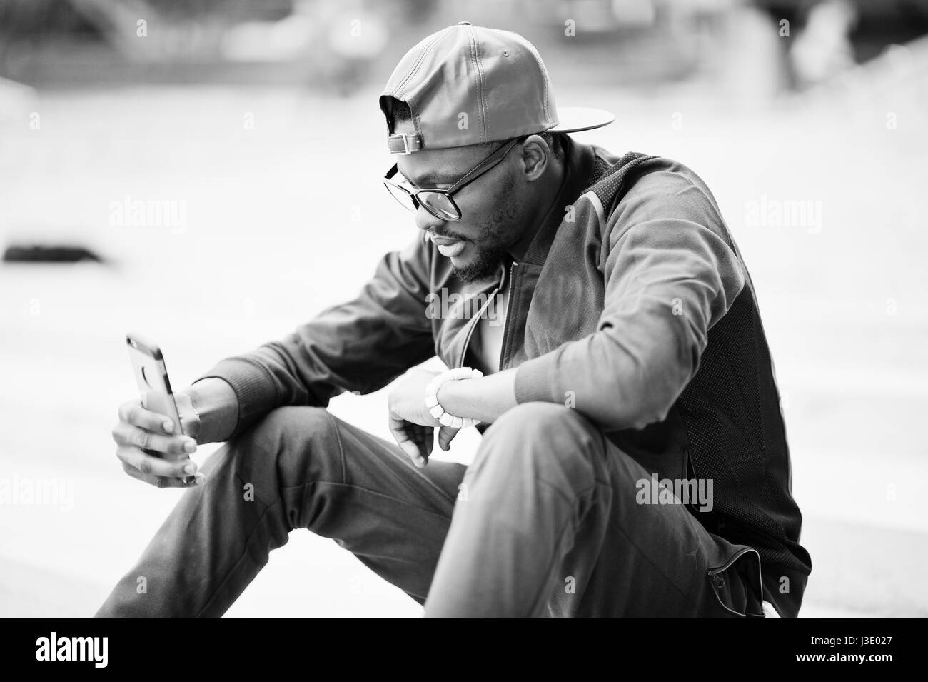 Ritratto di stile americano africano uomo su sportswear, cappello e occhiali seduti sulle scale con il telefono a portata di mano. Gli uomini neri modello street fashion. Foto Stock