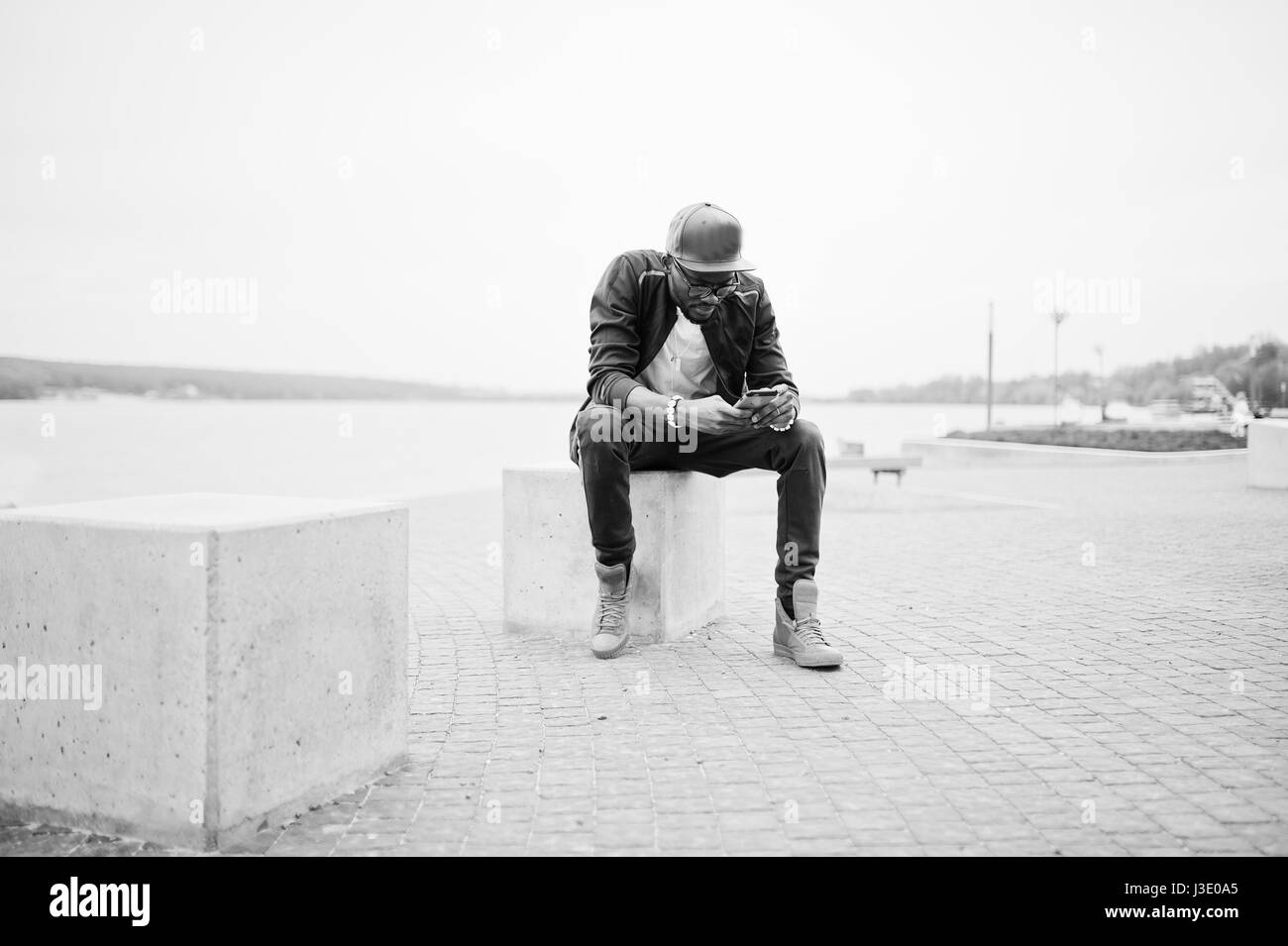Ritratto di stile americano africano uomo su sportswear, cappello e occhiali seduti al cubo di pietra con il telefono a portata di mano. Gli uomini neri modello street fashion. Foto Stock