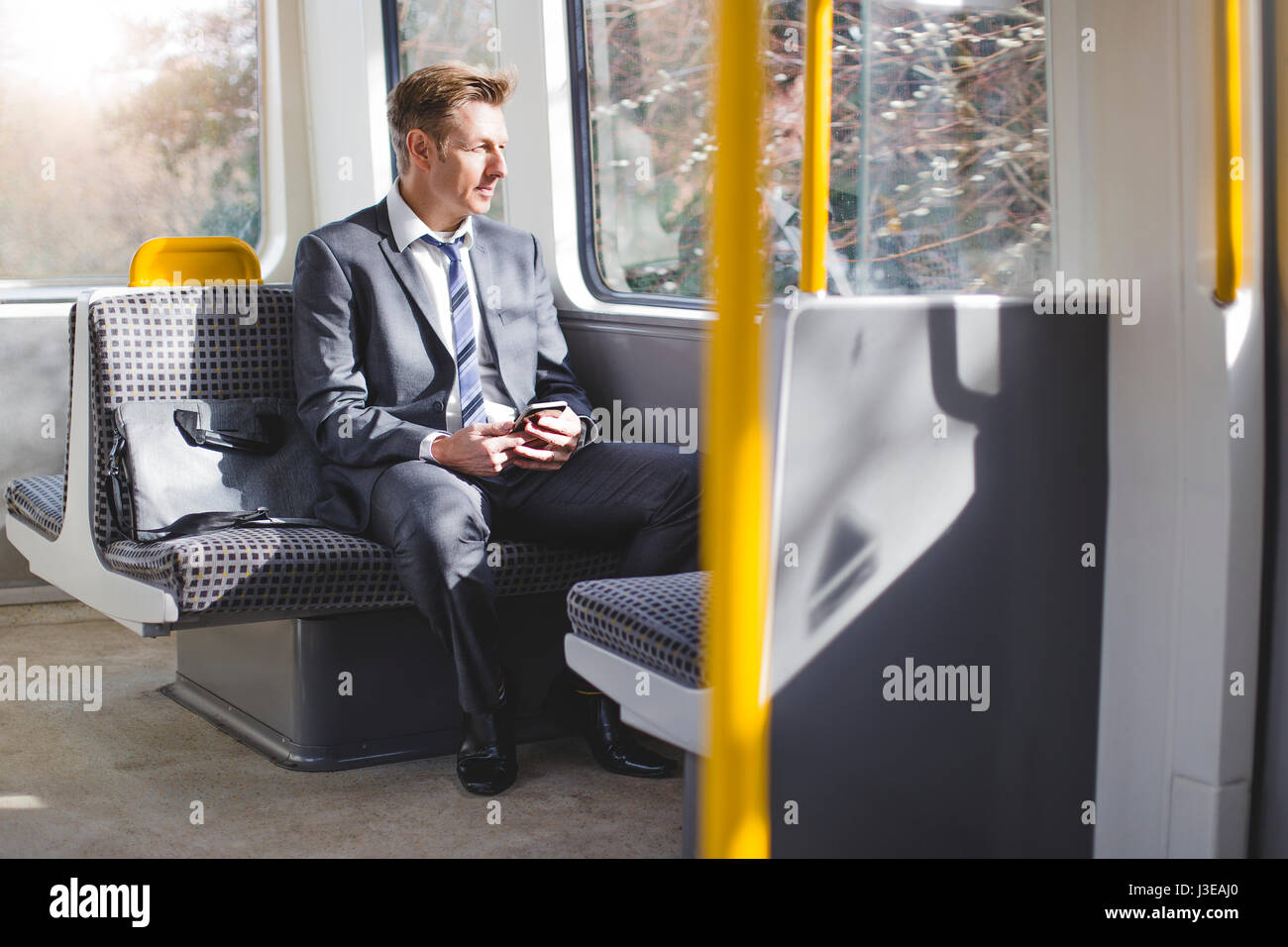 Business formale uomo seduto sul treno con la sua borsa. È lui a guardare fuori dalla finestra e ha un telefono intelligente nelle sue mani. Foto Stock