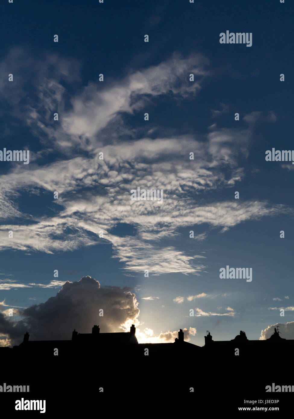 Tramonto dietro a profilarsi tetti con apertura a ventaglio di raggi ad un blu scuro cielo chiazzato Foto Stock