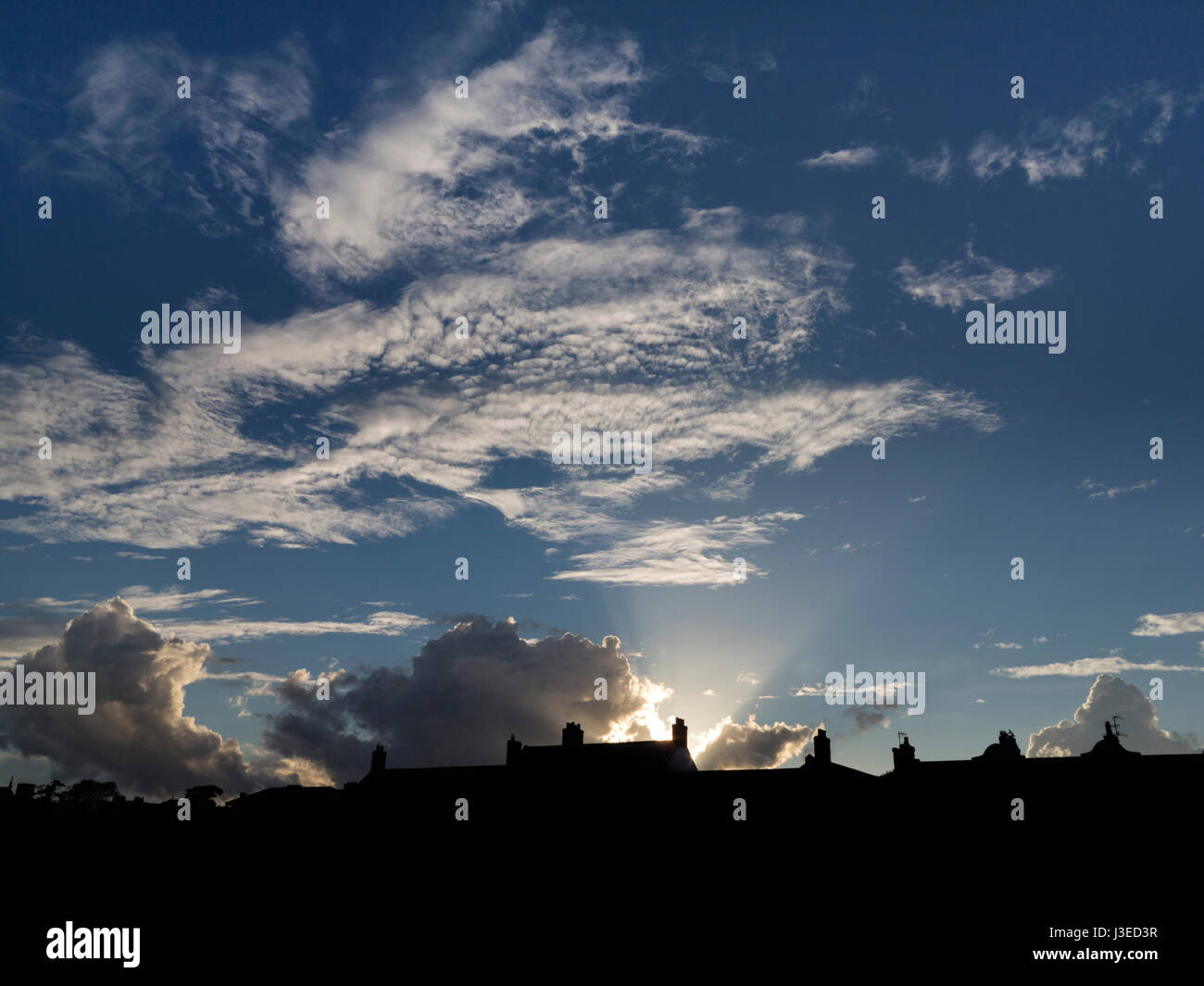 Tramonto dietro a profilarsi tetti con apertura a ventaglio di raggi ad un blu scuro cielo chiazzato Foto Stock