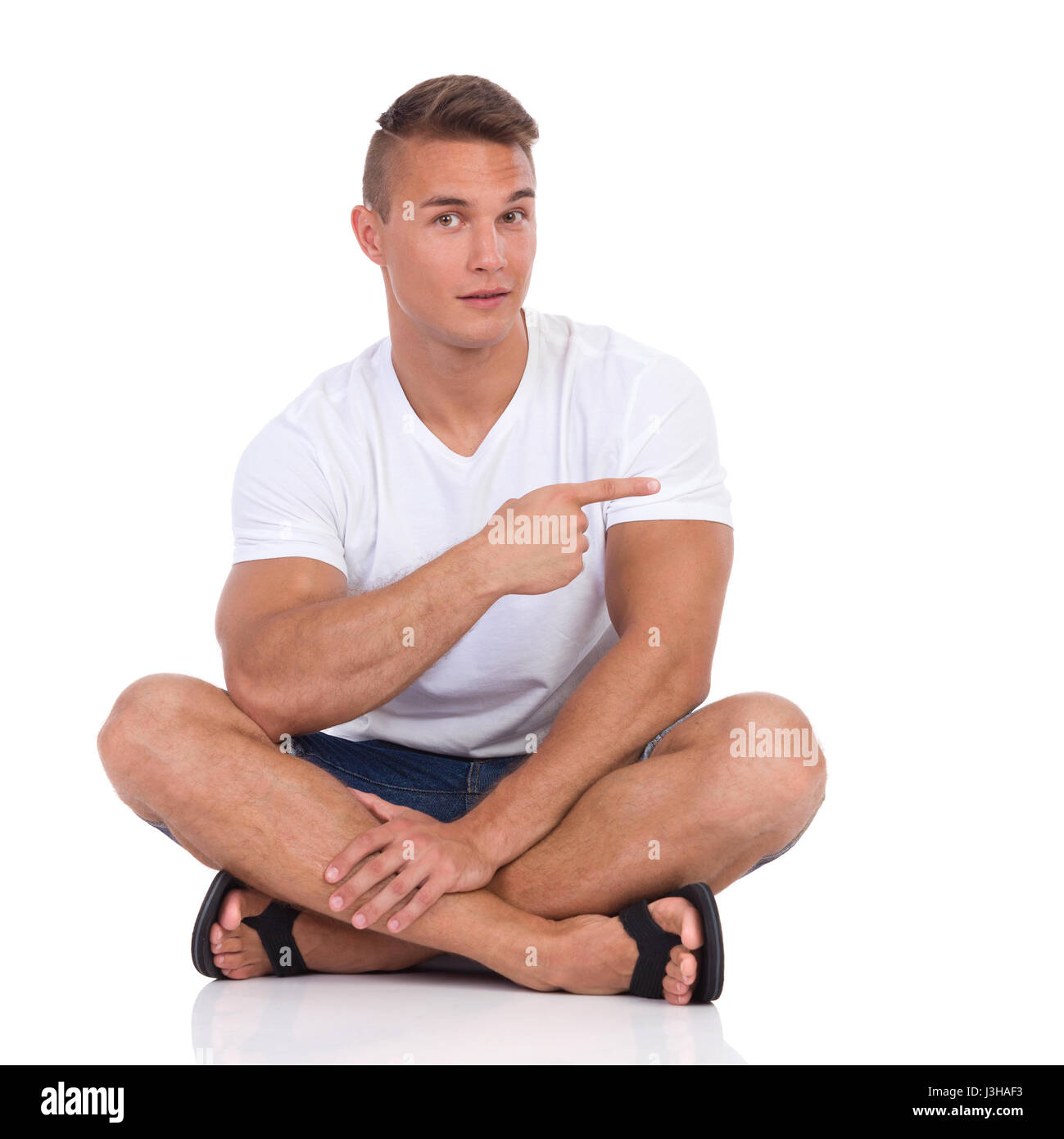 Bel Uomo in camicia bianca e jeans corti, seduto sul pavimento con le gambe  incrociate, puntamento e guardando la fotocamera. Lunghezza completa studio  shot isolato o Foto stock - Alamy