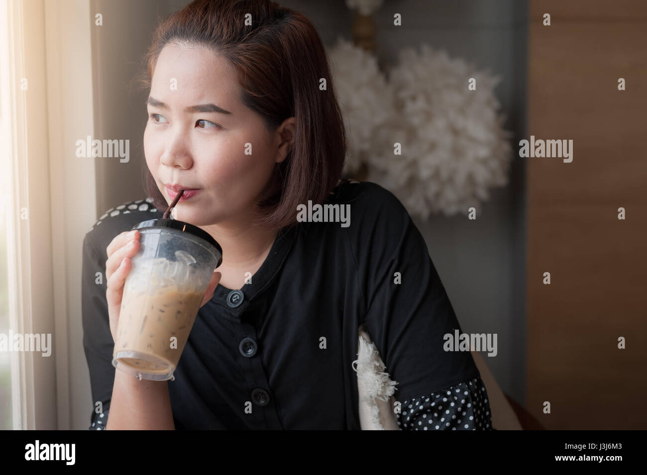 Giovane donna asiatica bere latte ghiacciata caffè accanto alla finestra in cafe con buon feeling. Fine Settimana lifestyle concetto astratto Foto Stock