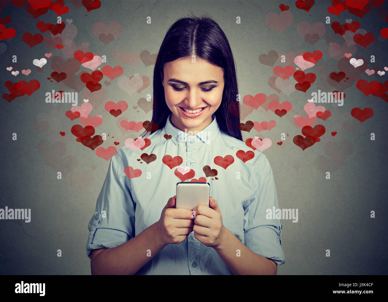 Bella donna felice amore invio messaggio di testo sul telefono cellulare con cuori rossi volare lontano dallo schermo isolato su sfondo grigio. Umani positivi em Foto Stock