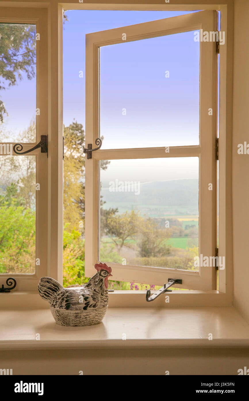 La luce del mattino che mostra attraverso un paese di lingua inglese cottage finestra dove un pollo di ceramica si siede a guardare la vista. Foto Stock