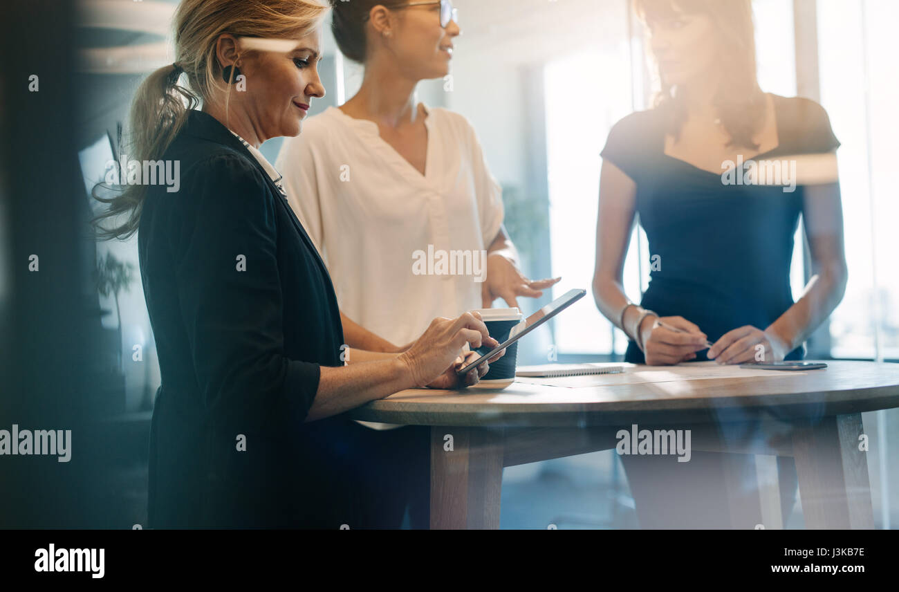 Tre colleghi femmina avente un incontro permanente intorno a un tavolo. Donna matura con tavoletta digitale con i colleghi per discutere in attesa. Foto Stock