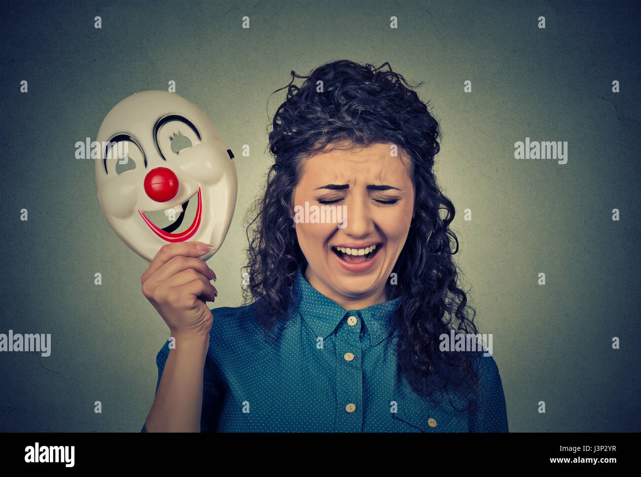 Ritratto giovane arrabbiato pianto urlando donna tenendo un clown maschera esprimere allegria felicità isolato sul muro grigio Sfondo. Le emozioni umane Foto Stock