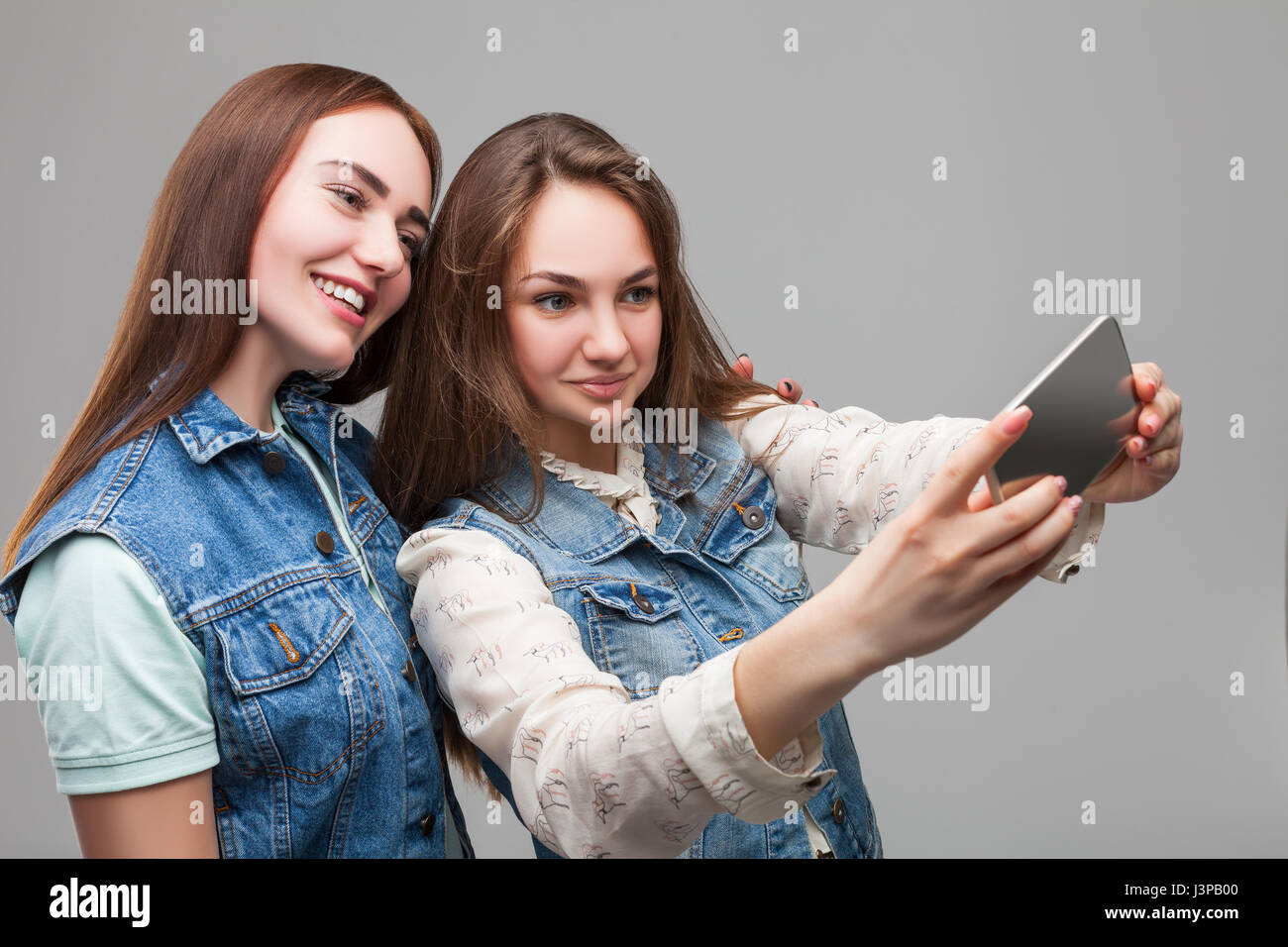 Due ragazze sorridenti in giacche denum rende selfie sulla fotocamera del telefono in studio. Amicizia femminile. Tempo libero di felice ragazze Foto Stock