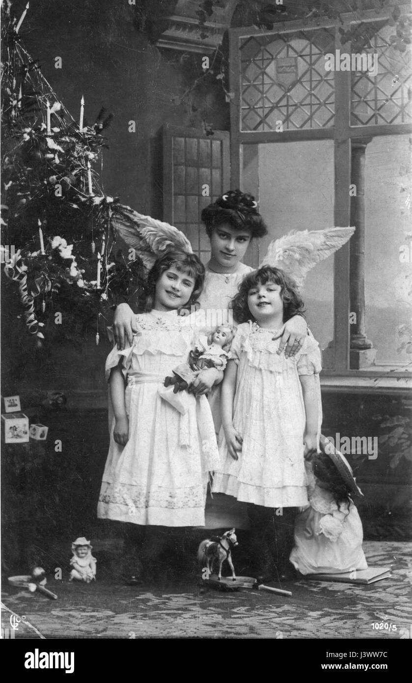 Una madre vestita come un Angelo, con due ragazze, probabilmente le sue figlie, c. 1910. Si trovano in un albero di Natale decorato con candele accese, strisce di carta a spirale e corde. Le ragazze tengono nuove bambole, e anche visibile sono un cavallo giocattolo, blocchi giocattolo e badminton giocattolo (?) racchette. 1910. Per vedere le mie altre immagini d'epoca legate al Natale, Cerca: Prestor vintage Holiday Foto Stock