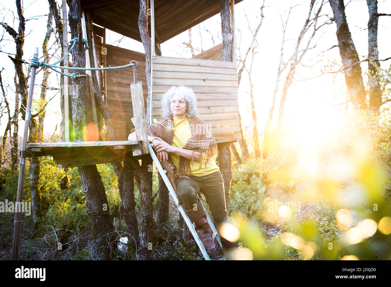 Donna matura appoggiata contro woodland treehouse scaletta al tramonto Foto Stock