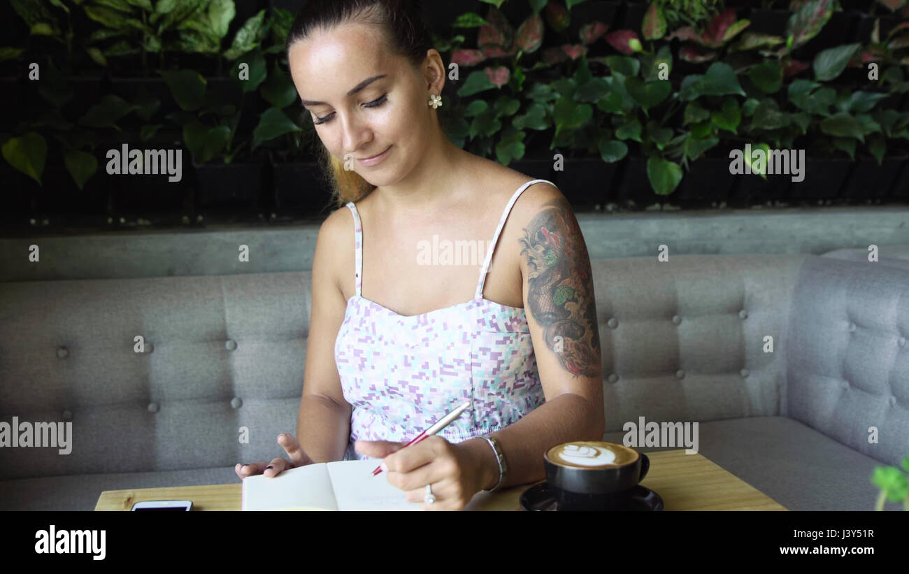 Giovane donna prety con tattoo iscritto nel suo diario, diario. Modello caucasico con capelli lunghi. Tazza di caffè e lo smartphone sul tavolo, Foto Stock