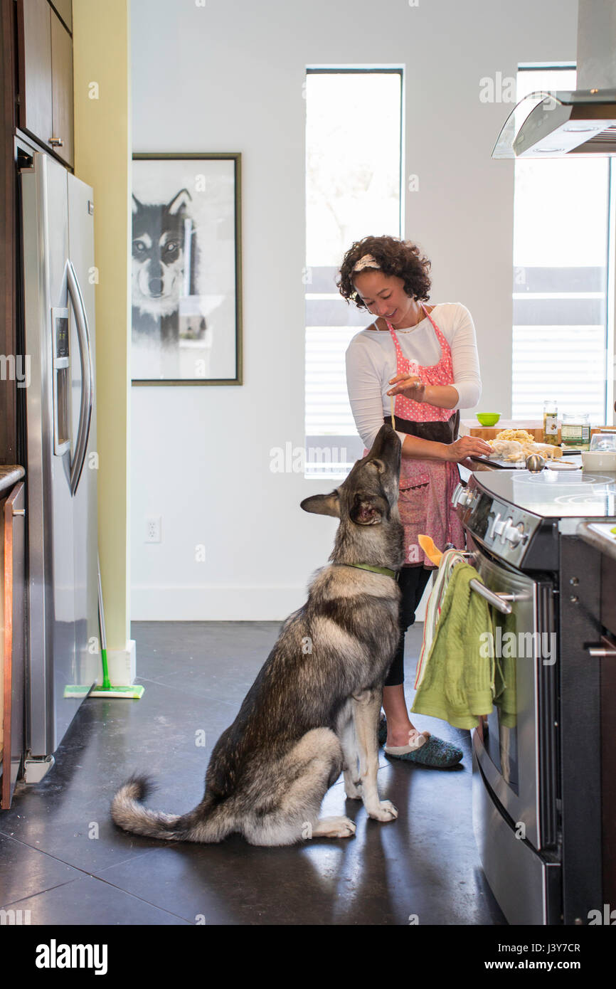 Donna matura per la cottura in cucina, alimentando il suo cane la pasta a base di noodle Foto Stock