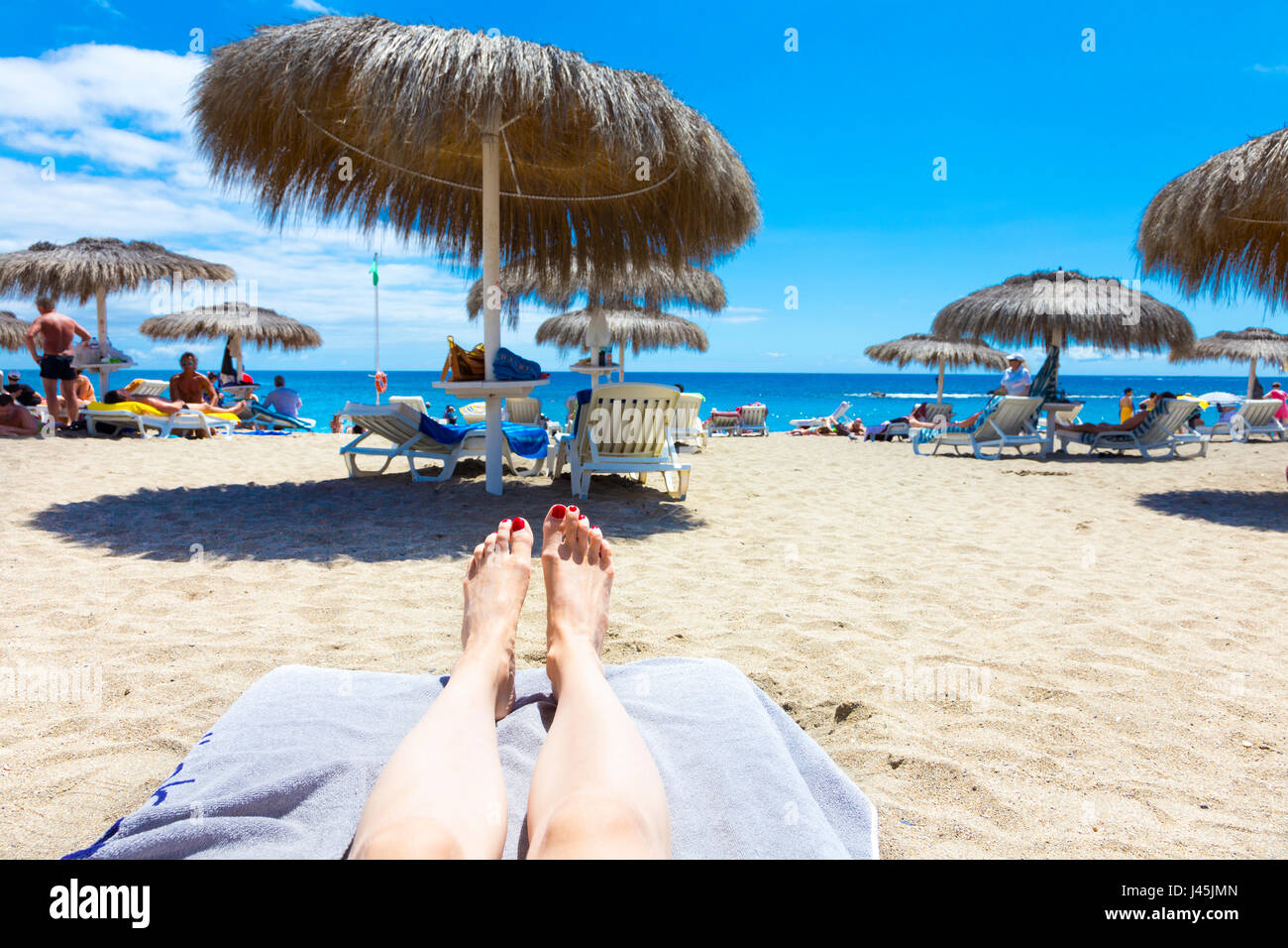 Gambe e piedi appoggiati su una sedia a sdraio con ombrelloni in background a Bahia Del Duque Beach a Tenerife, Spagna Foto Stock