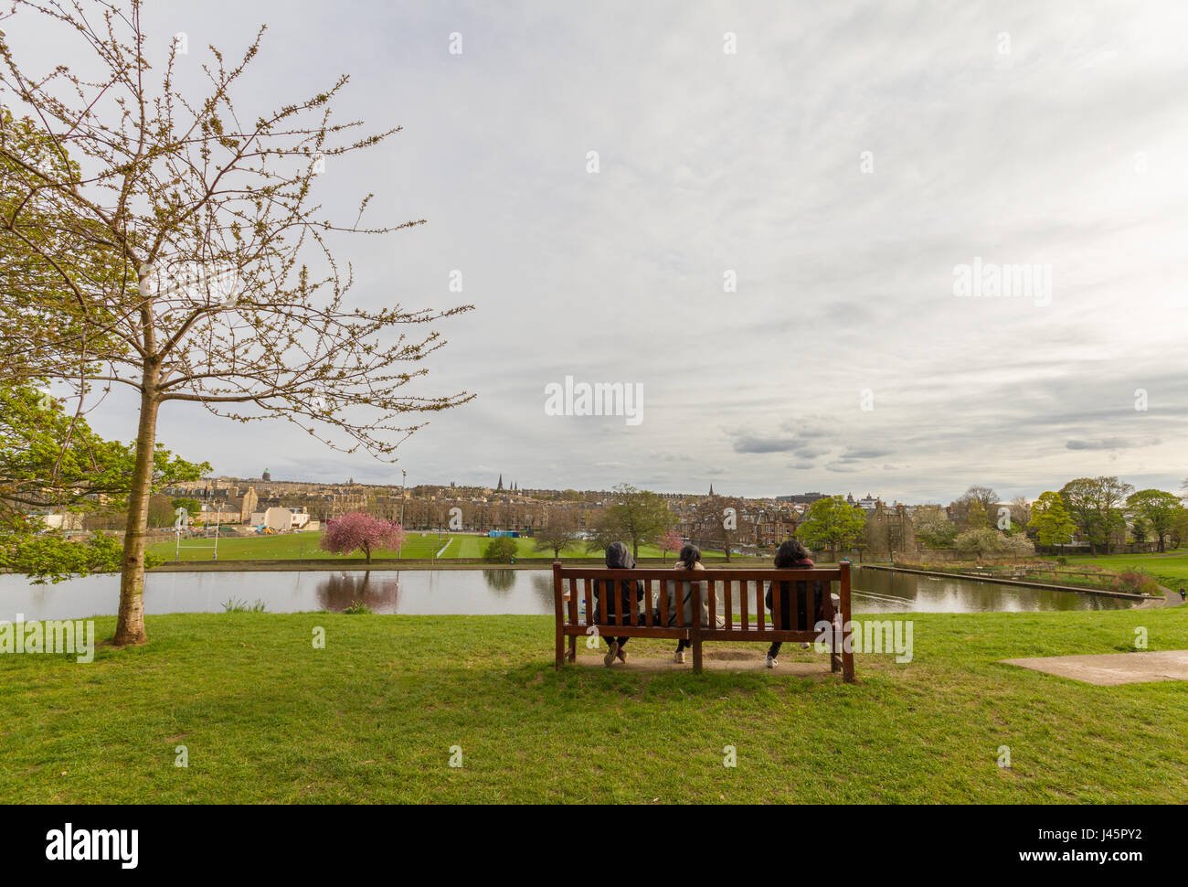 Romantica vista di tre persone sedute su una panchina guardando verso lo stagno a Inverleith Park di Edimburgo, Scozia, su un parzialmente nuvoloso dopo a molla Foto Stock