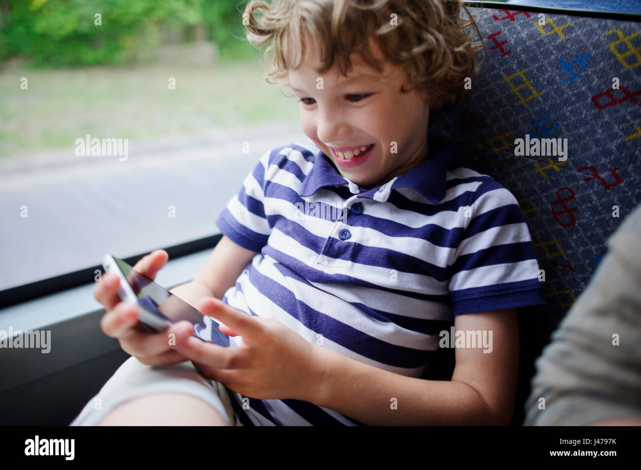 Un piccolo passeggero di un autobus di città con uno smartphone a portata di mano. Il ragazzo guarda lo schermo dello smartphone e allegramente sorrisi. Foto Stock