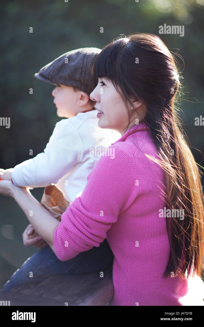 Giovane e bella madre asiatica con lunghi capelli neri che indossa un rosa maglione cashmere tenendo il suo figlio in un marrone newsboy cap come la luce del sole i suoi capelli Foto Stock