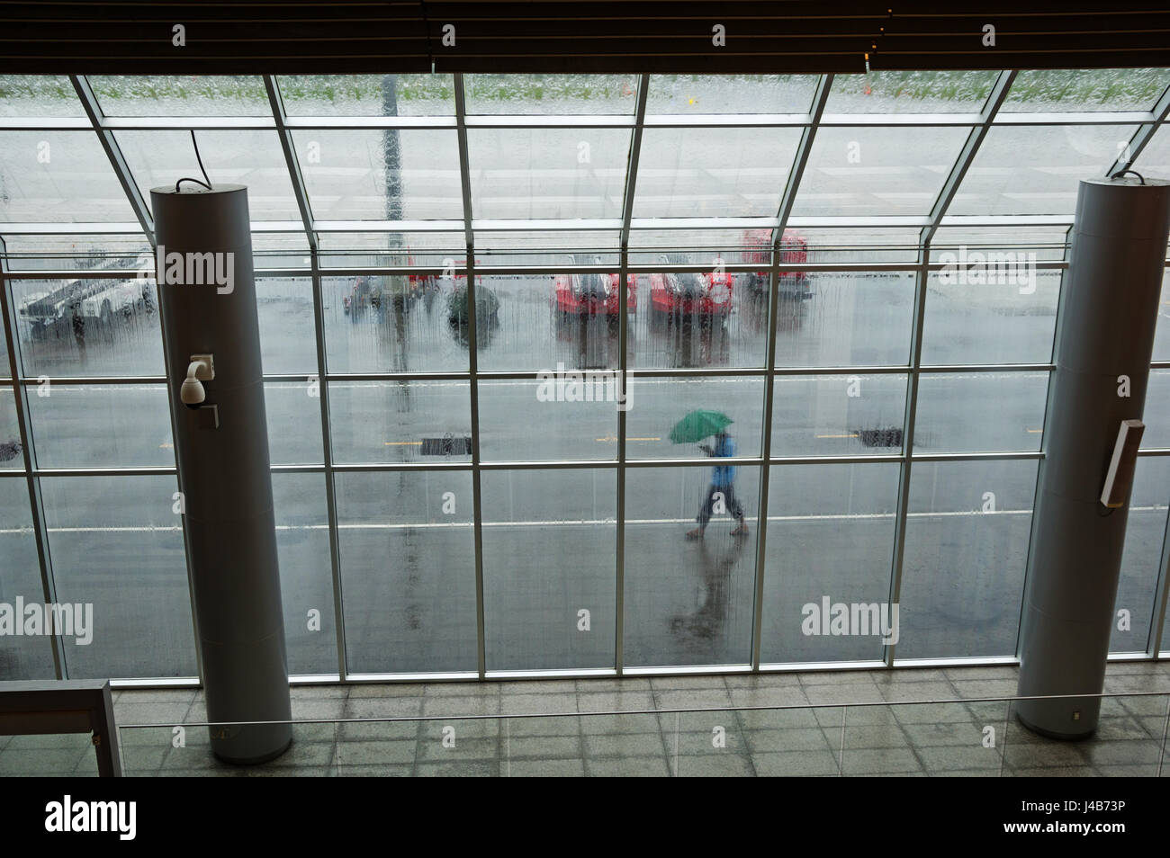 Guardando fuori attraverso una finestra di airport in un giorno di pioggia con una persona che cammina da esterno con ombrellone Foto Stock