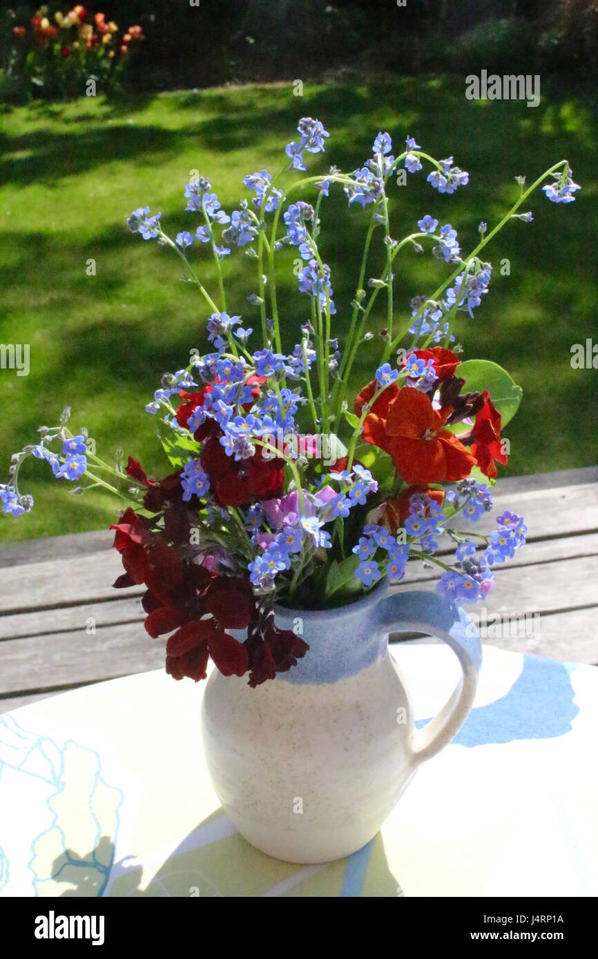 Posy di piccoli fiori in blu e crema Caraffa in ceramica seduta sul tavolo con coperchio con giardino in background Foto Stock