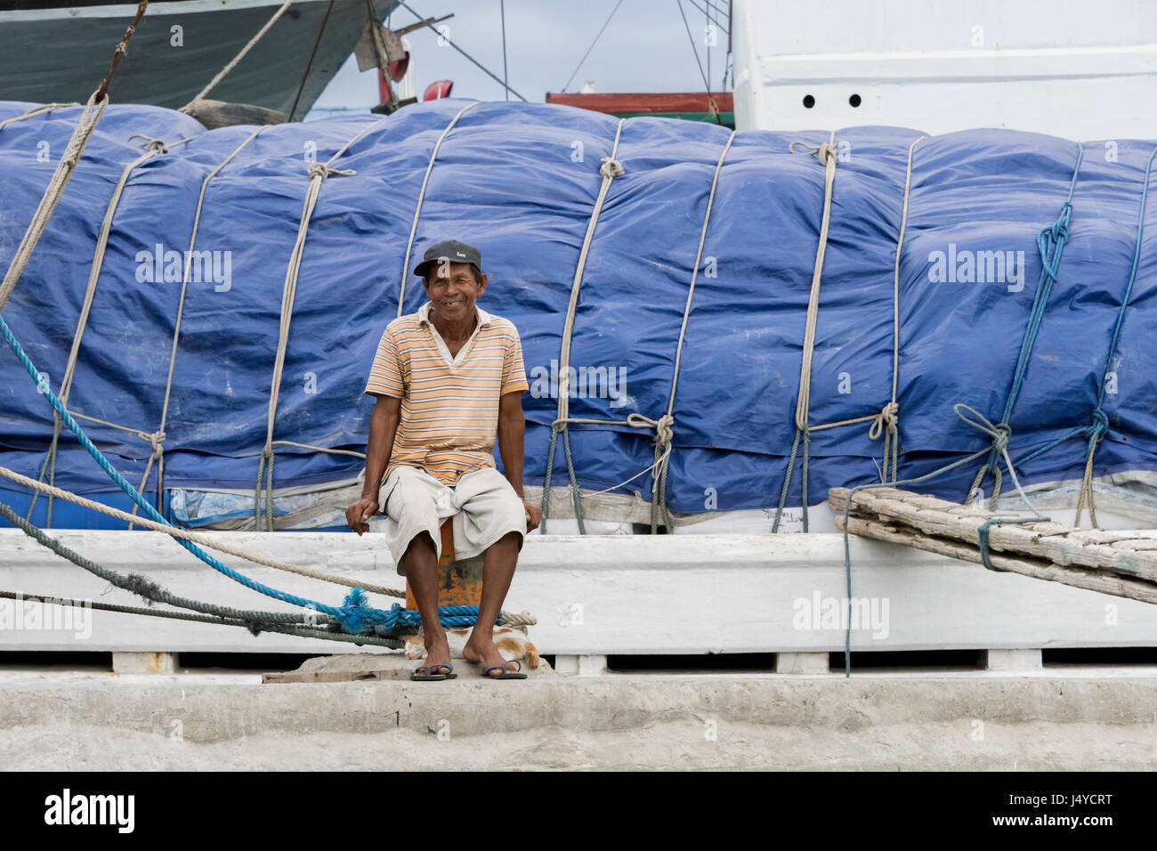 Marinaio sorridente seduto su un bollard con il suo gatto pelo ai suoi piedi, accanto a un pinisi, il Porto di Sunda Kelapa, Jakarta, Indonesia Foto Stock