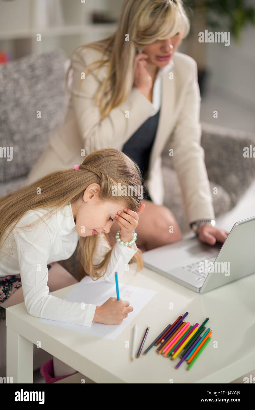 Donna che utilizza il telefono in ufficio a casa con il computer portatile mentre la ragazza giovane disegno Foto Stock
