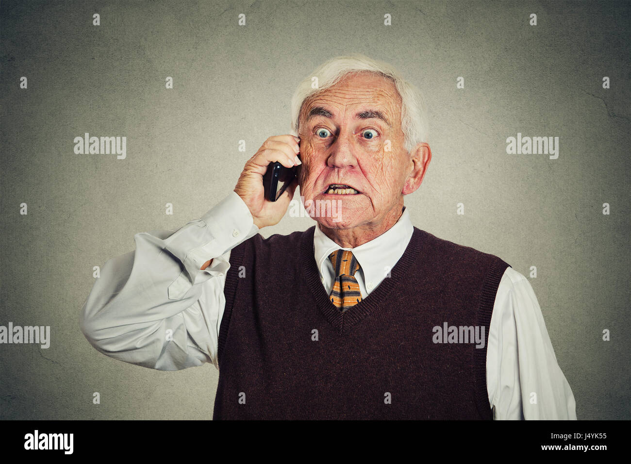 Arrabbiato senior uomo parlando al telefono cellulare isolata sul muro grigio Sfondo. le emozioni negative Foto Stock