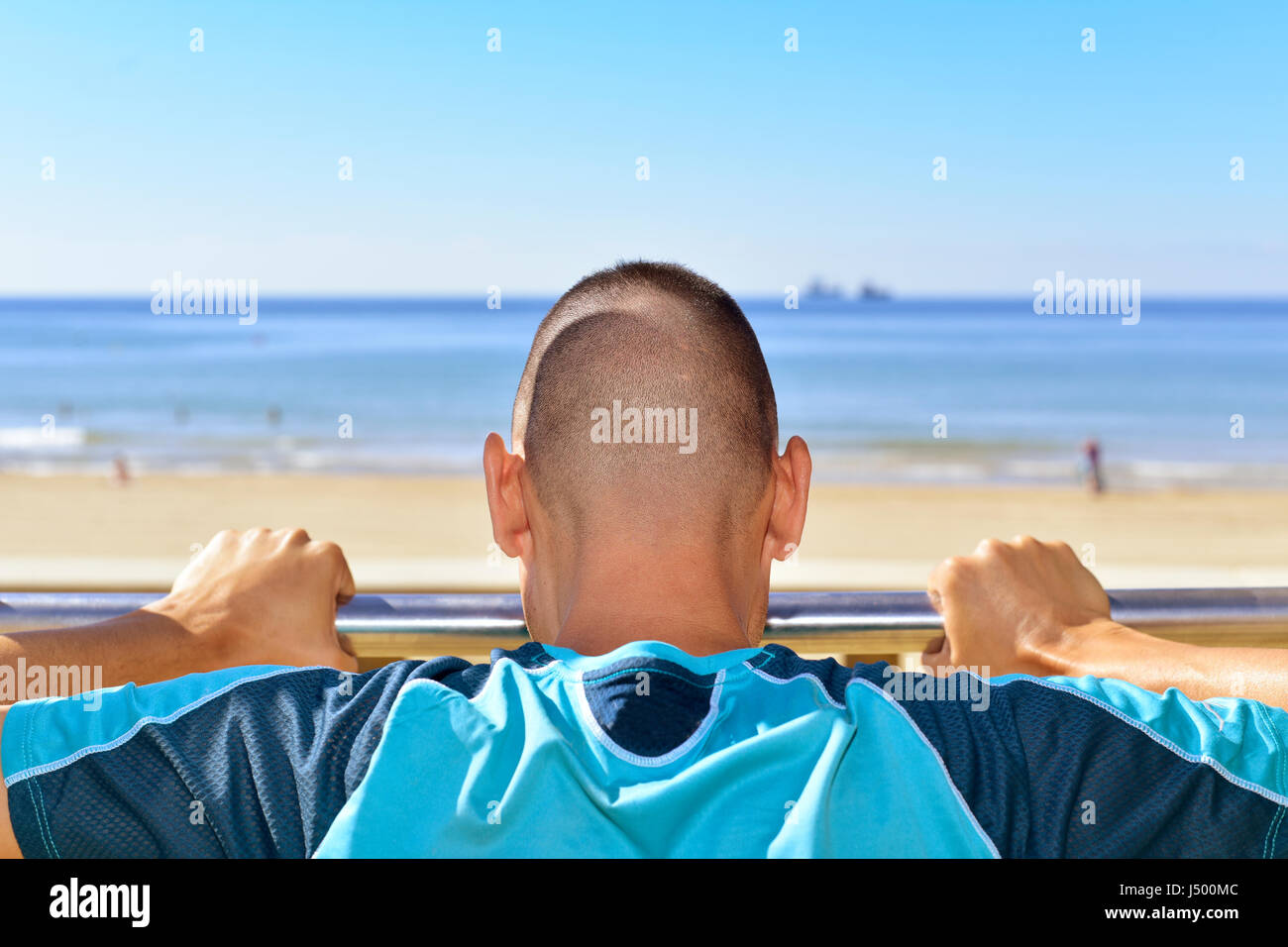 Primo piano di un giovane sportivo caucasici in sport abbigliamento visto da dietro facendo inclinare push-up in una barra metallica all'aperto, accanto al mare Foto Stock