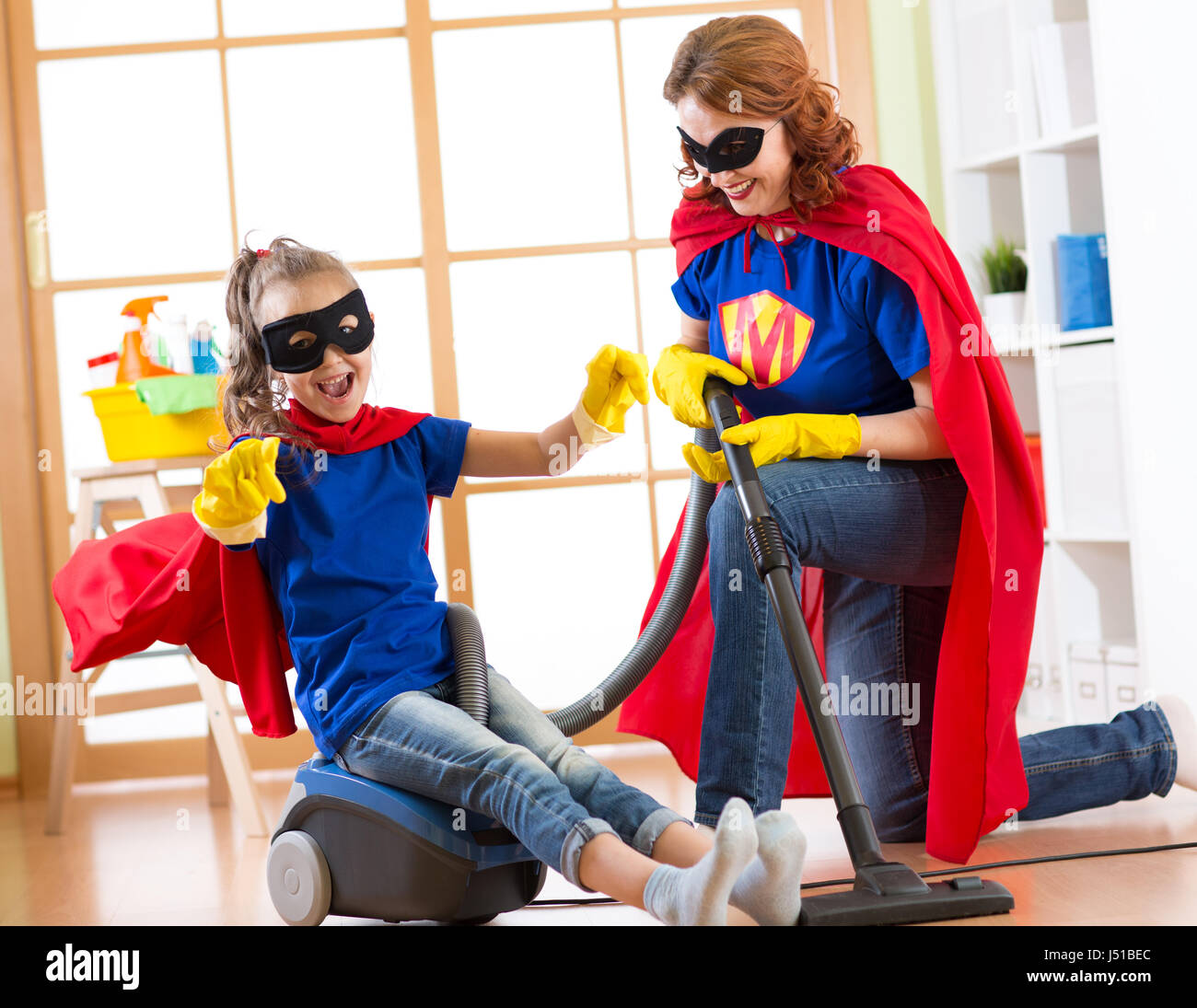 Il bambino e la madre vestita come supereroi utilizzando aspirapolvere in camera. Famiglia di mezza età della donna e la figlia hanno un divertimento durante la pulizia del pavimento. Foto Stock