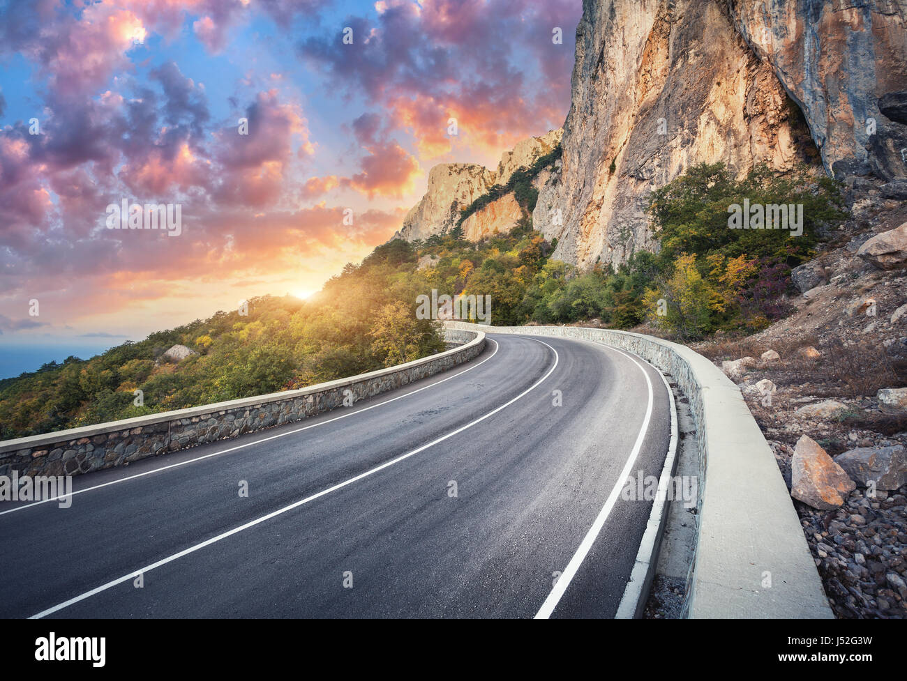 Bella strada asfaltata. Colorato paesaggio con alte rocce, strada di montagna con un asfalto perfetto, alberi e soleggiata incredibile cielo nuvoloso al tramonto in summ Foto Stock