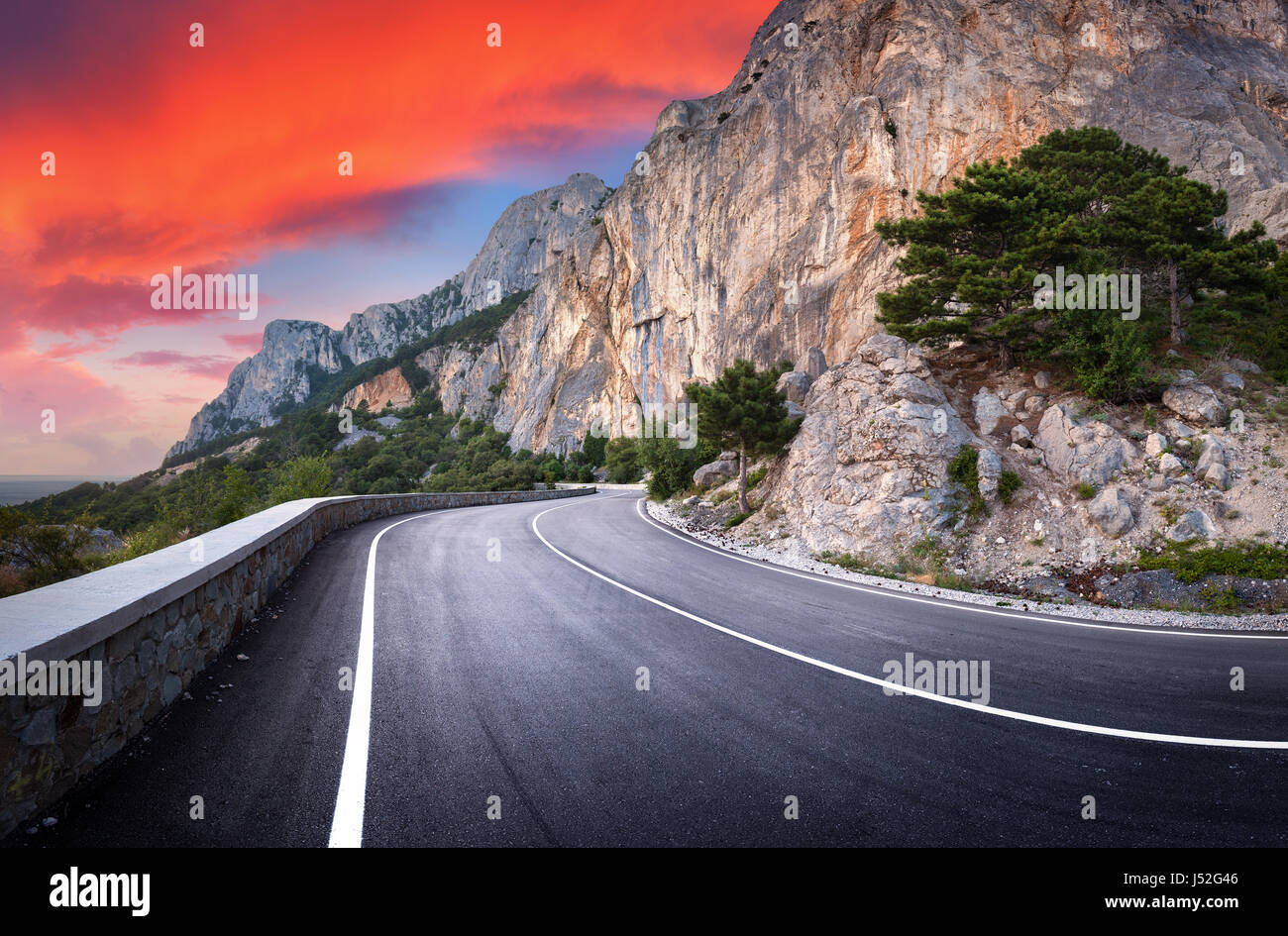 Strada asfaltata. Paesaggio con una bella strada di montagna con un asfalto perfetto, alte rocce, alberi, incredibile cielo rosso con le nuvole al tramonto Foto Stock