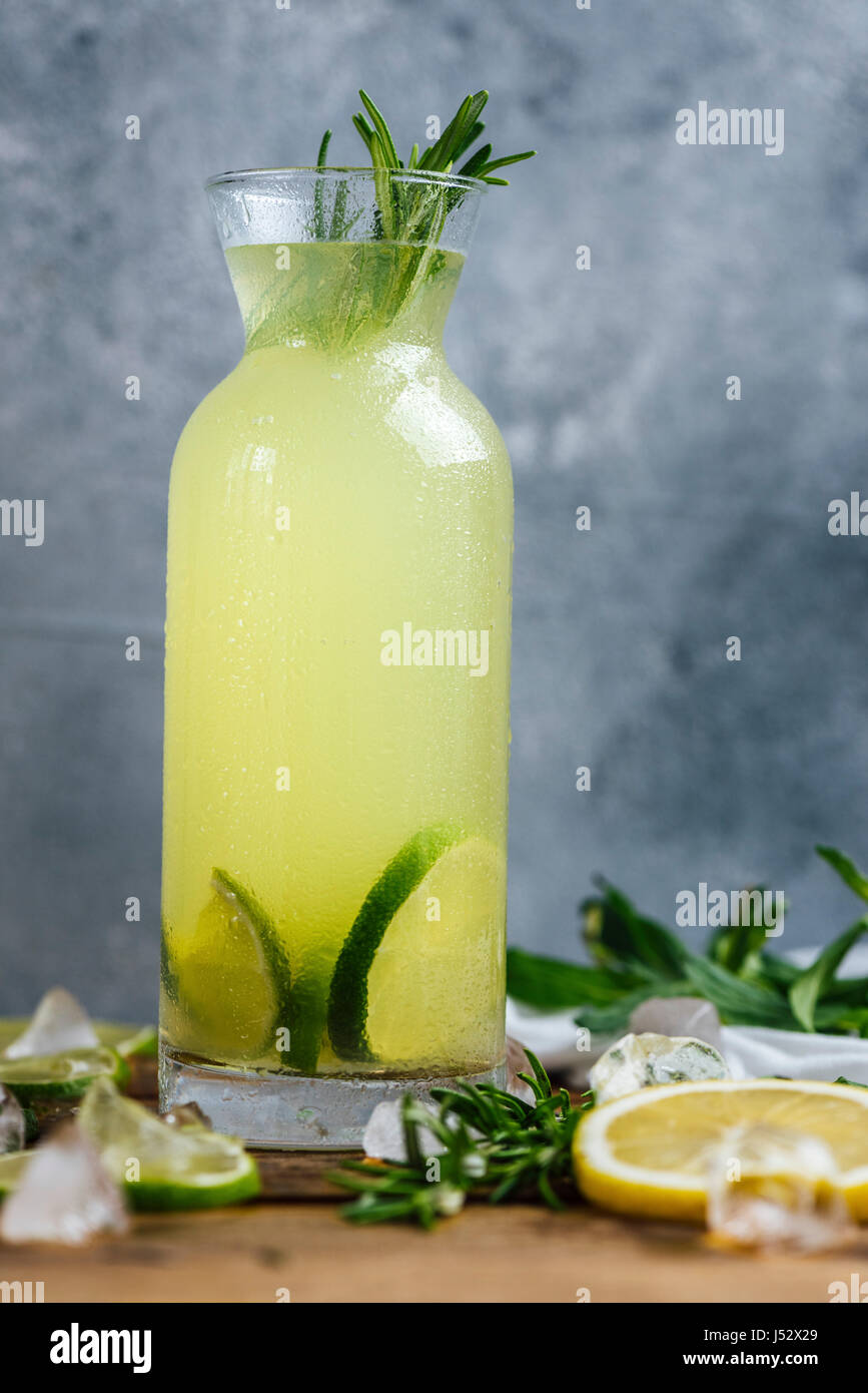 La limonata aromatizzata con menta e rosmarino servito in bottiglia fotografato su una tavola di legno con uno sfondo grigio dalla vista frontale. Fette di limone, calce Foto Stock