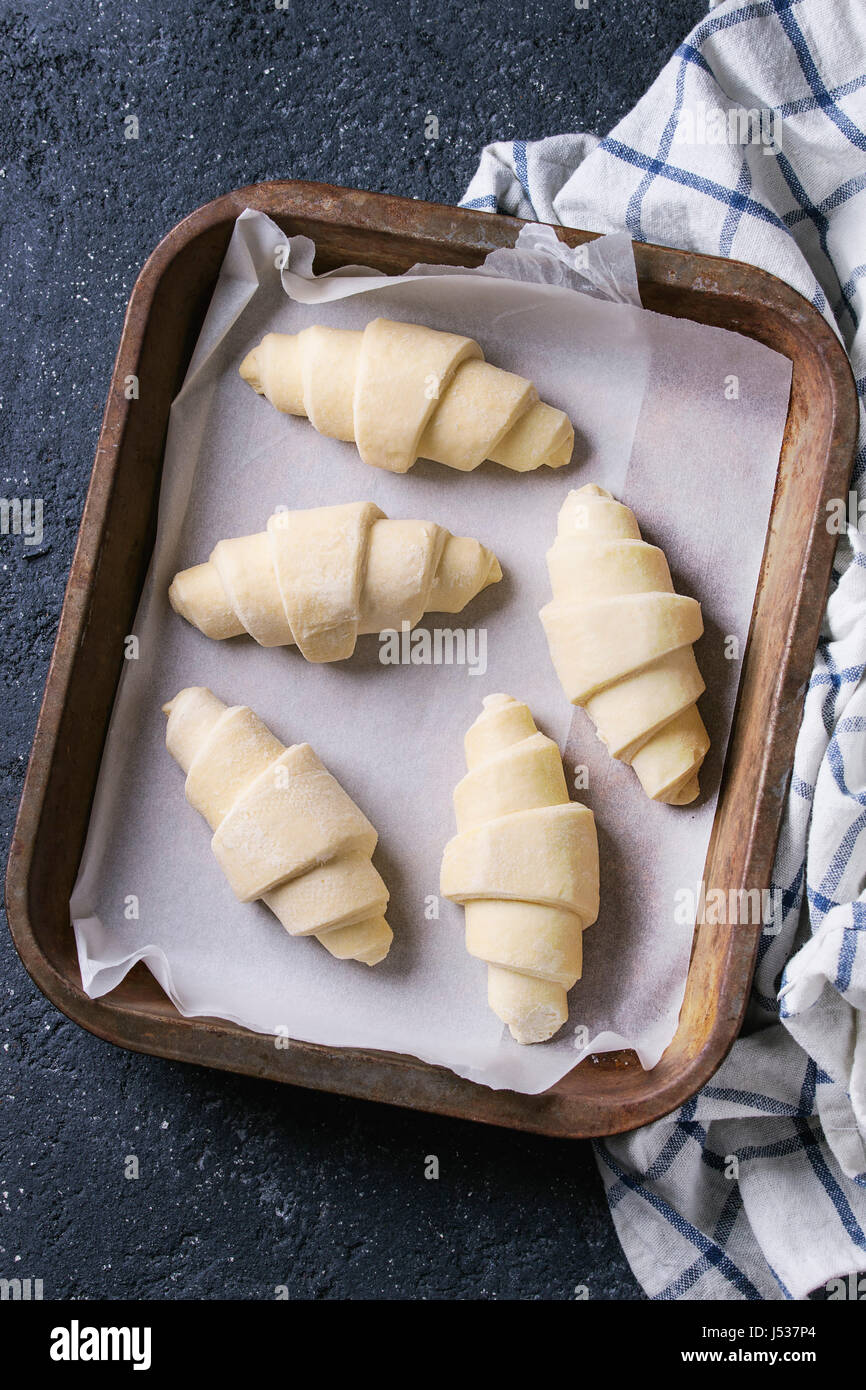 Materie crudi croissant sulla carta da forno nel vecchio forno il vassoio con carta asciugatutto per black texture di sfondo di calcestruzzo. Vista superiore Foto Stock