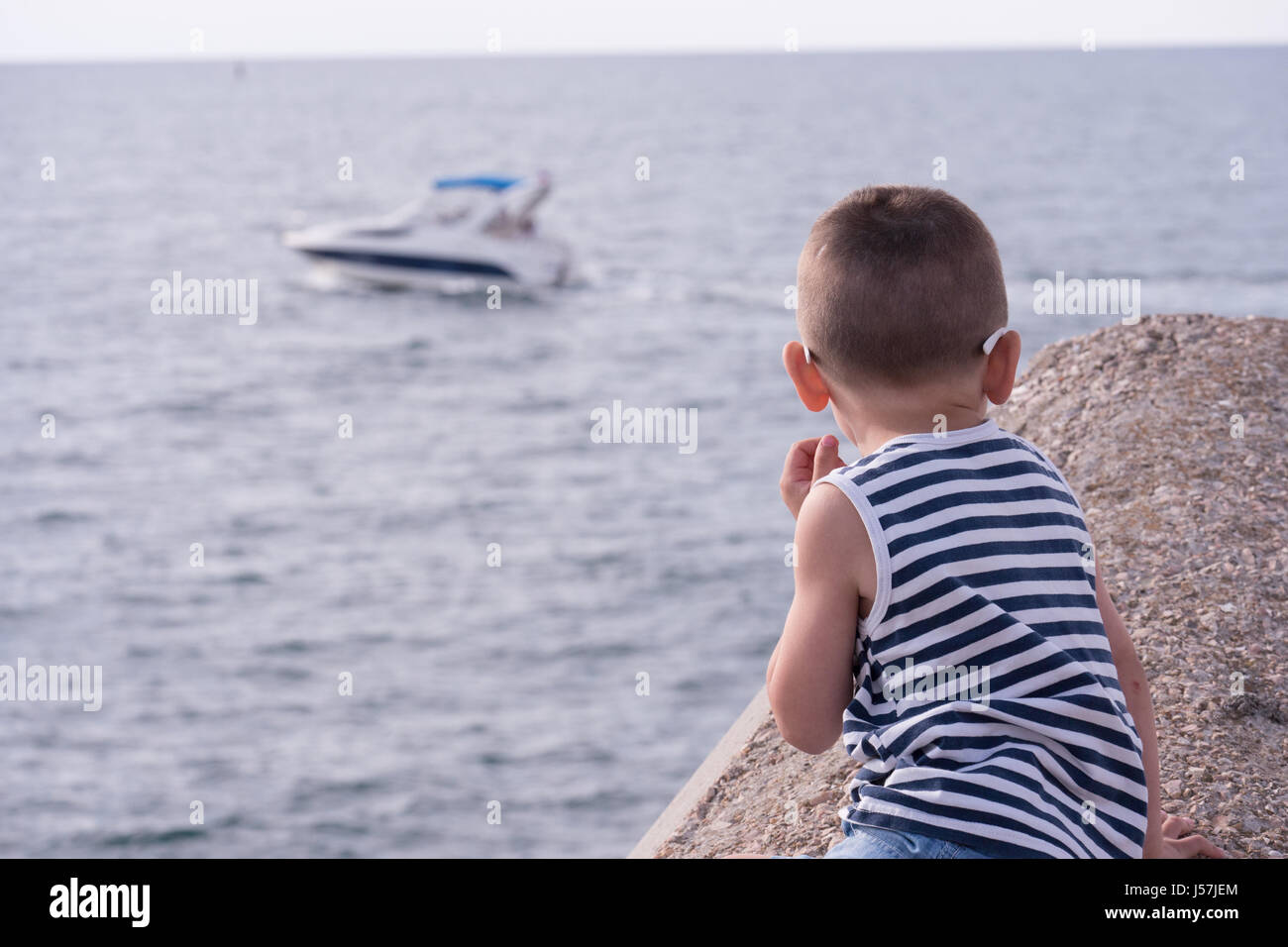 Ragazzino con gli occhiali da sole e giubbotto di striping guardando in lontananza sul mare con barca flottante Foto Stock