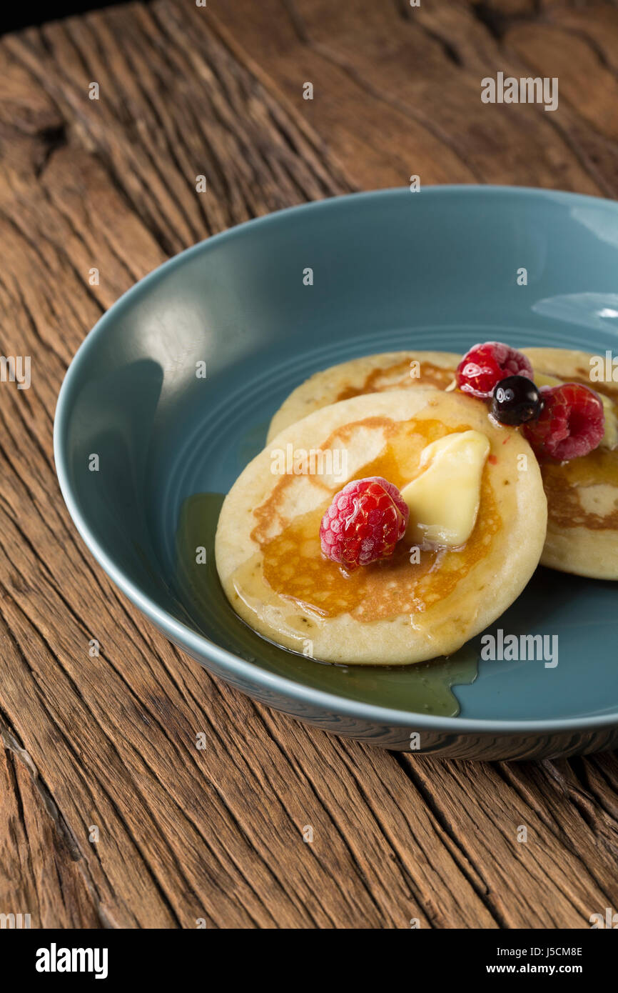 Tre i pancake freschi, una spruzzata di sciroppo e di frutti di bosco in una ciotola blu, seduto su di una tavola in legno rustico. Foto Stock