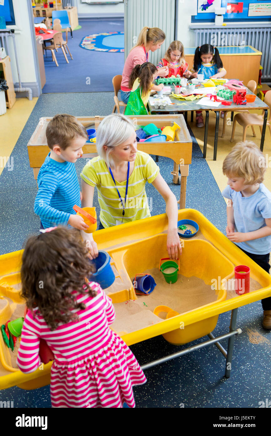 Vista in elevazione degli insegnanti delle attività con i bambini in vivaio. Alcuni stanno giocando in una scatola di sabbia e gli altri sono l'artigianato. Foto Stock