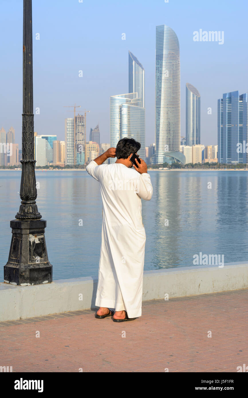 Vista dello Skyline di Abu Dhabi con un uomo vestito in tradizionali abiti arabi utilizzando un telefono cellulare, Emirati arabi uniti (EAU), Medio Oriente Foto Stock