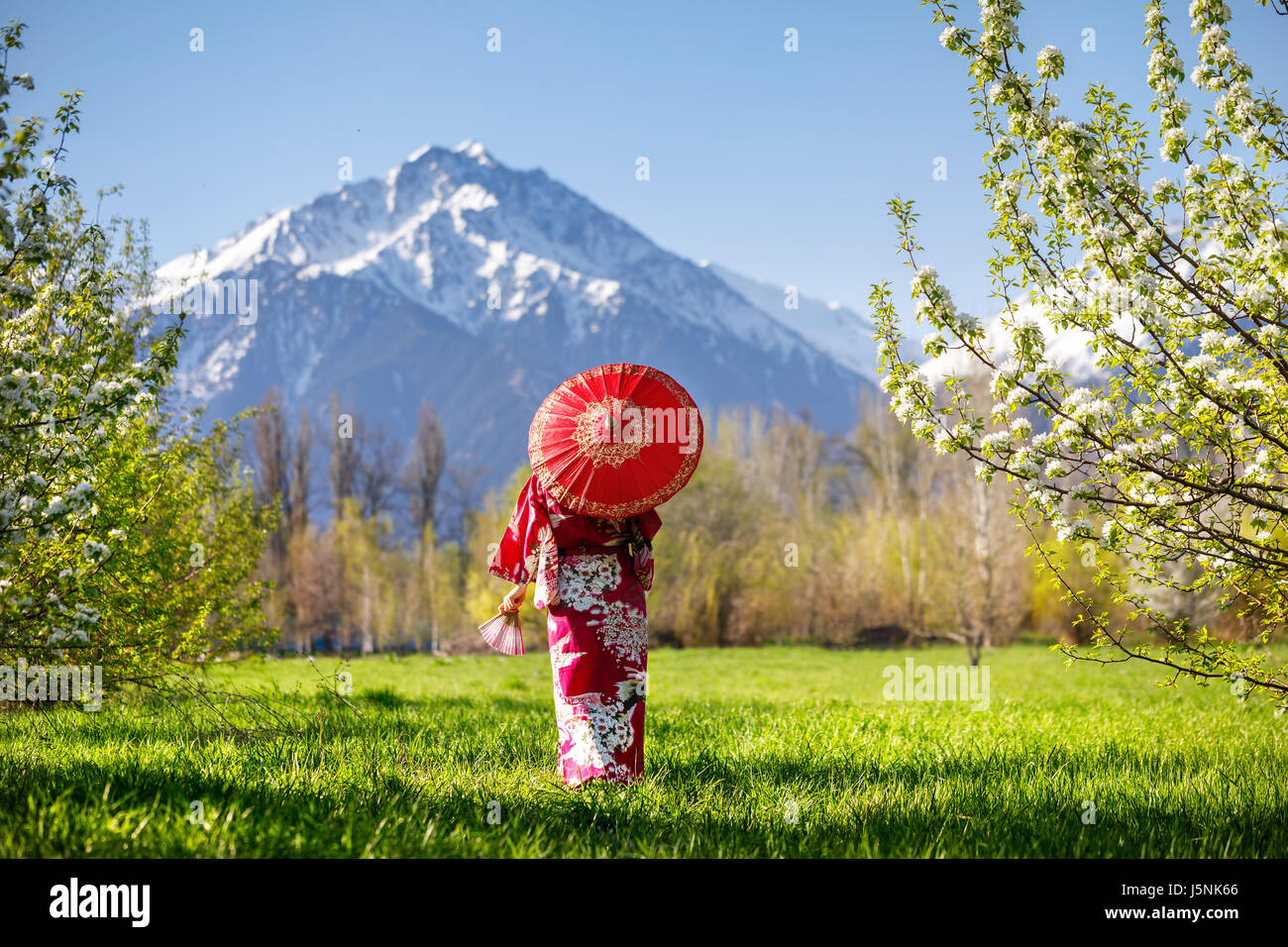 Donna in kimono con ombrellone rosso nel giardino con fiori di ciliegio a sfondo di montagna Foto Stock