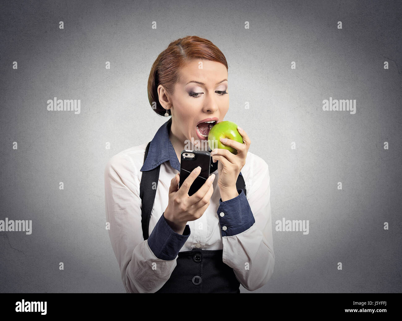 Closeup ritratto grave corporate business donna, deal maker per leggere le notizie su smart telefono cellulare azienda mangiare mela verde isolato muro grigio backgro Foto Stock