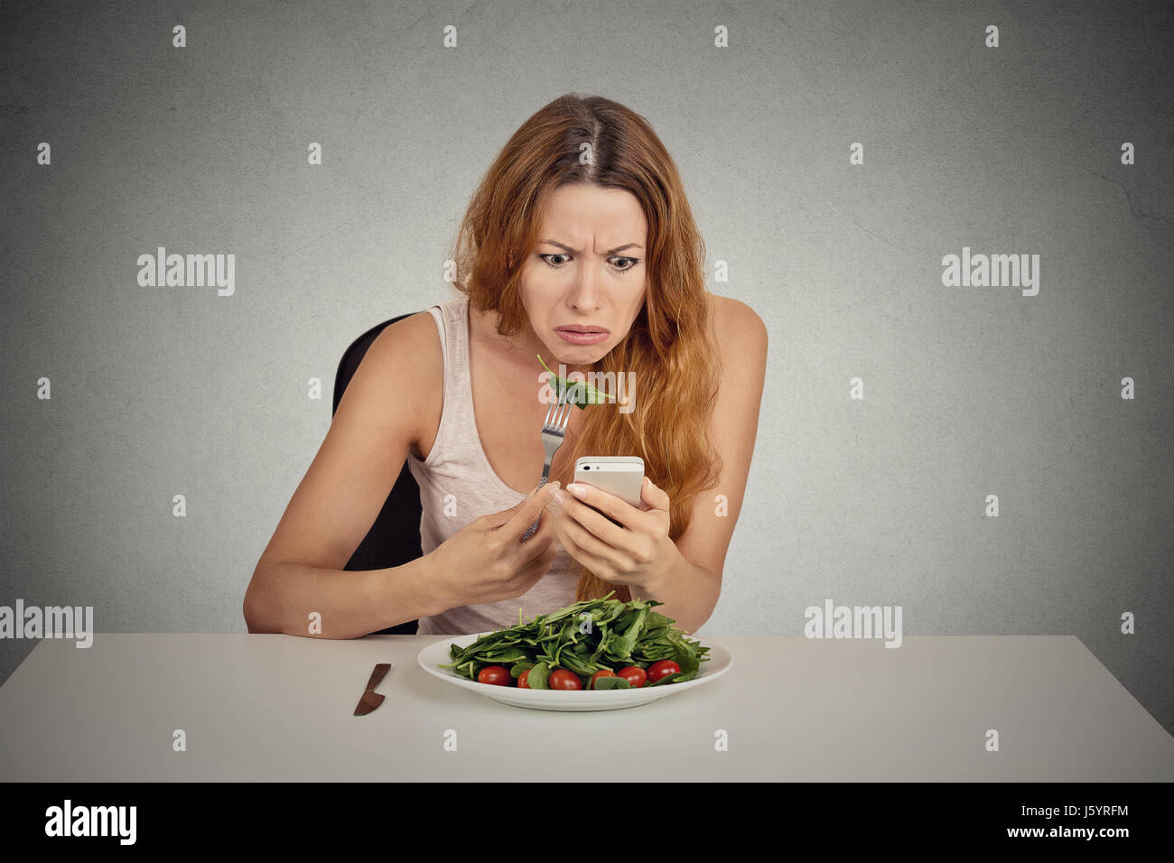 Ritratto ragazza giovane mangiare insalata verde guardando phone vedendo le cattive notizie o foto infastiditi confuso deluso faccia espressione isolato sul muro grigio Foto Stock